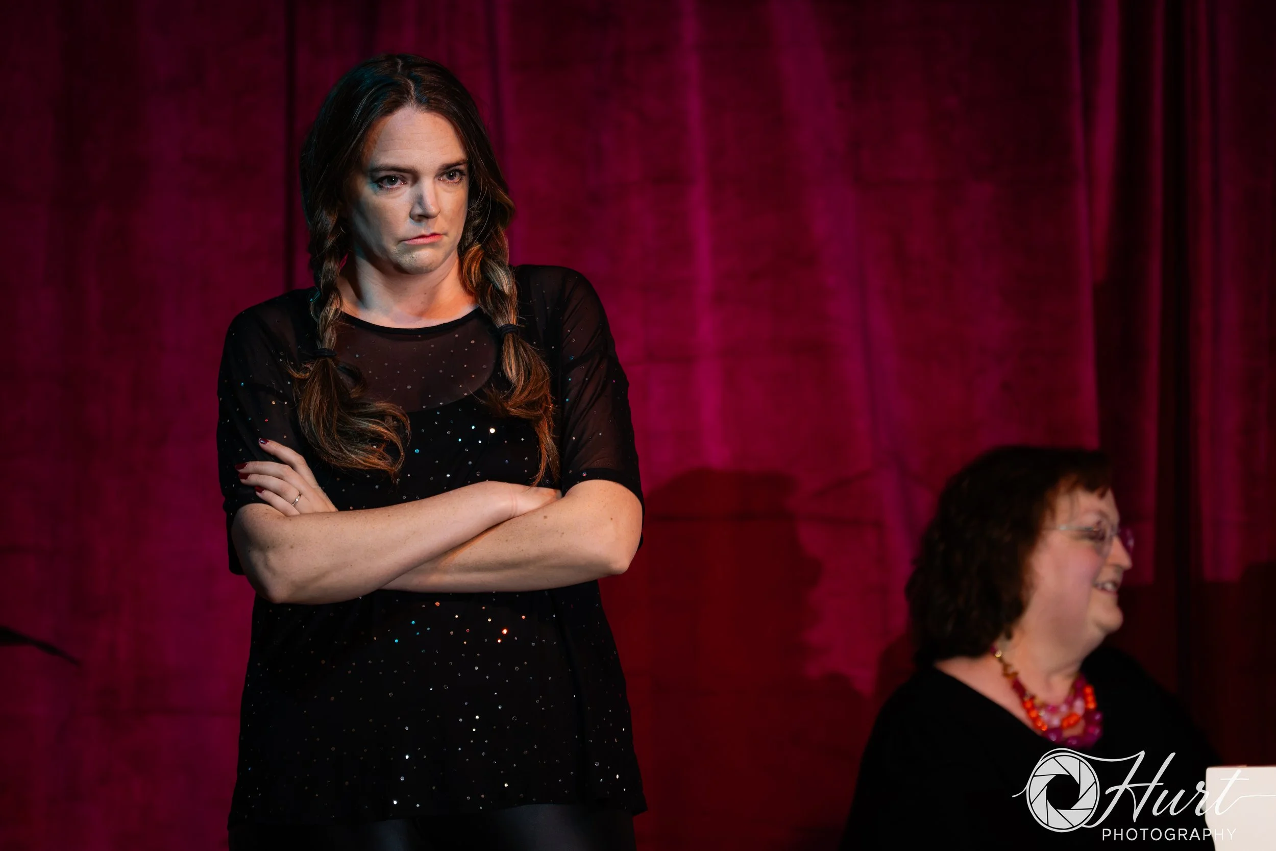 A woman with braids and a black dress with sparkles standing with her arms crossed in front of a magenta curtain.