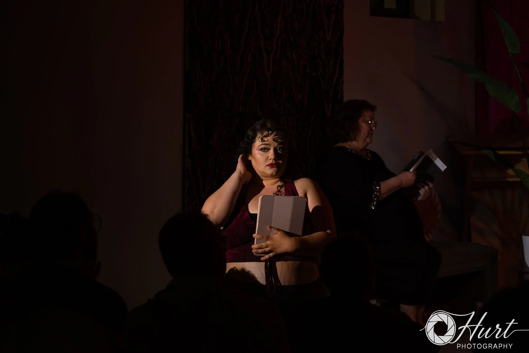 A woman in a black dress holds a tablet in the spotlight on stage. 