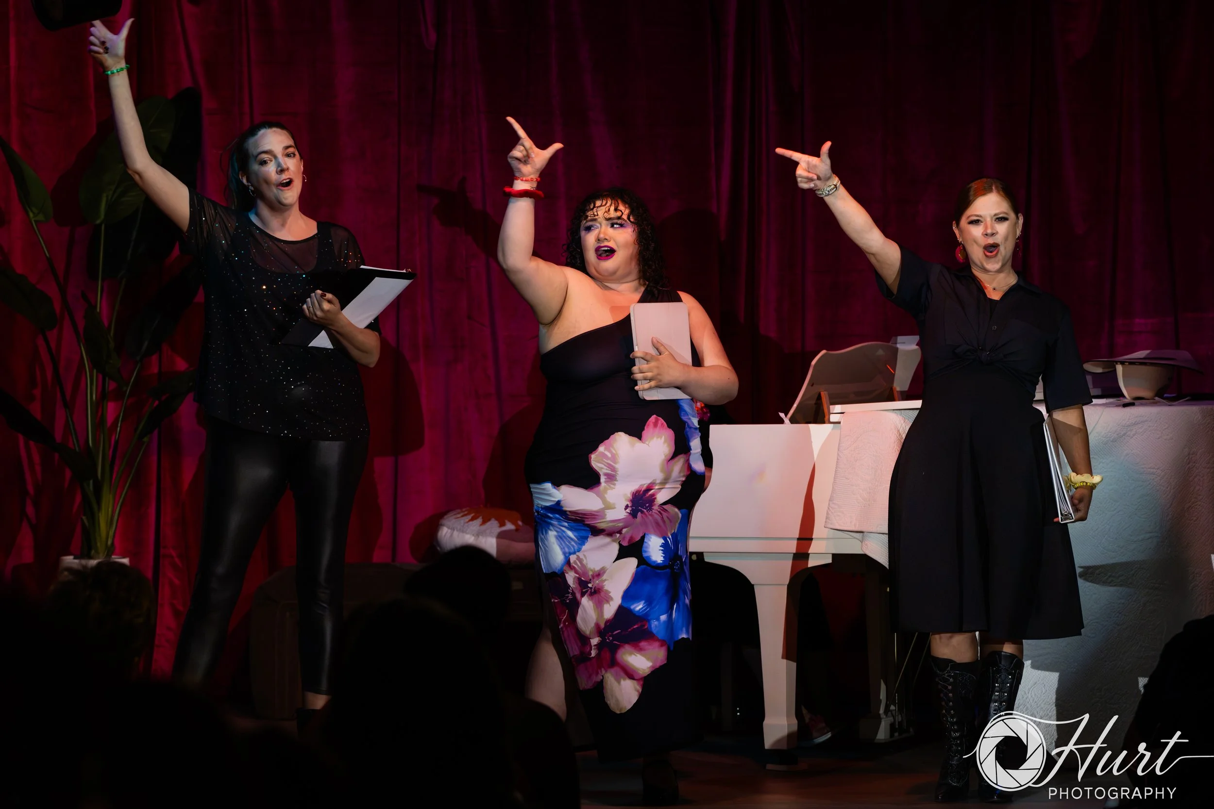 Three women performing on stage in front of a magenta curtain with their right arms raised in the air. 