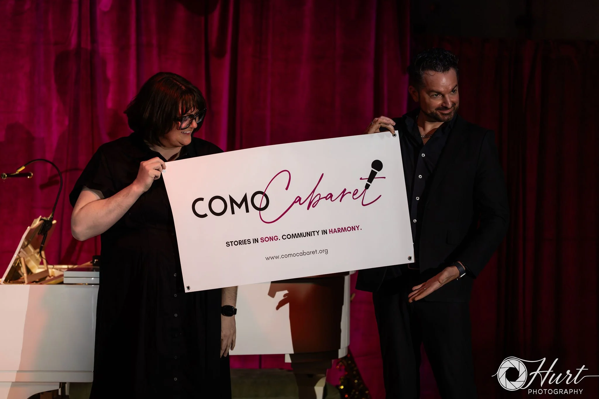 Two people holding a large sign that reads "COMO Cabaret" on a stage with magenta curtains, with a woman smiling and a man smiling and looking to the side.