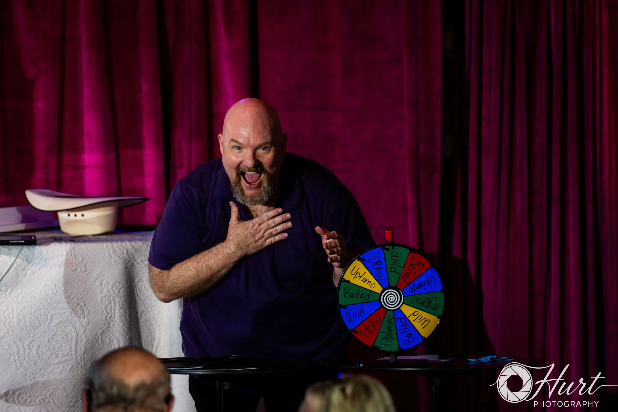 A man with a bald head and beard, wearing a dark shirt, is laughing and touching his chest with his right hand, sitting at a table with a colorful spinning wheel labeled with words like "WILD," "Ballad," and "Uptempo." A red curtain and a white table