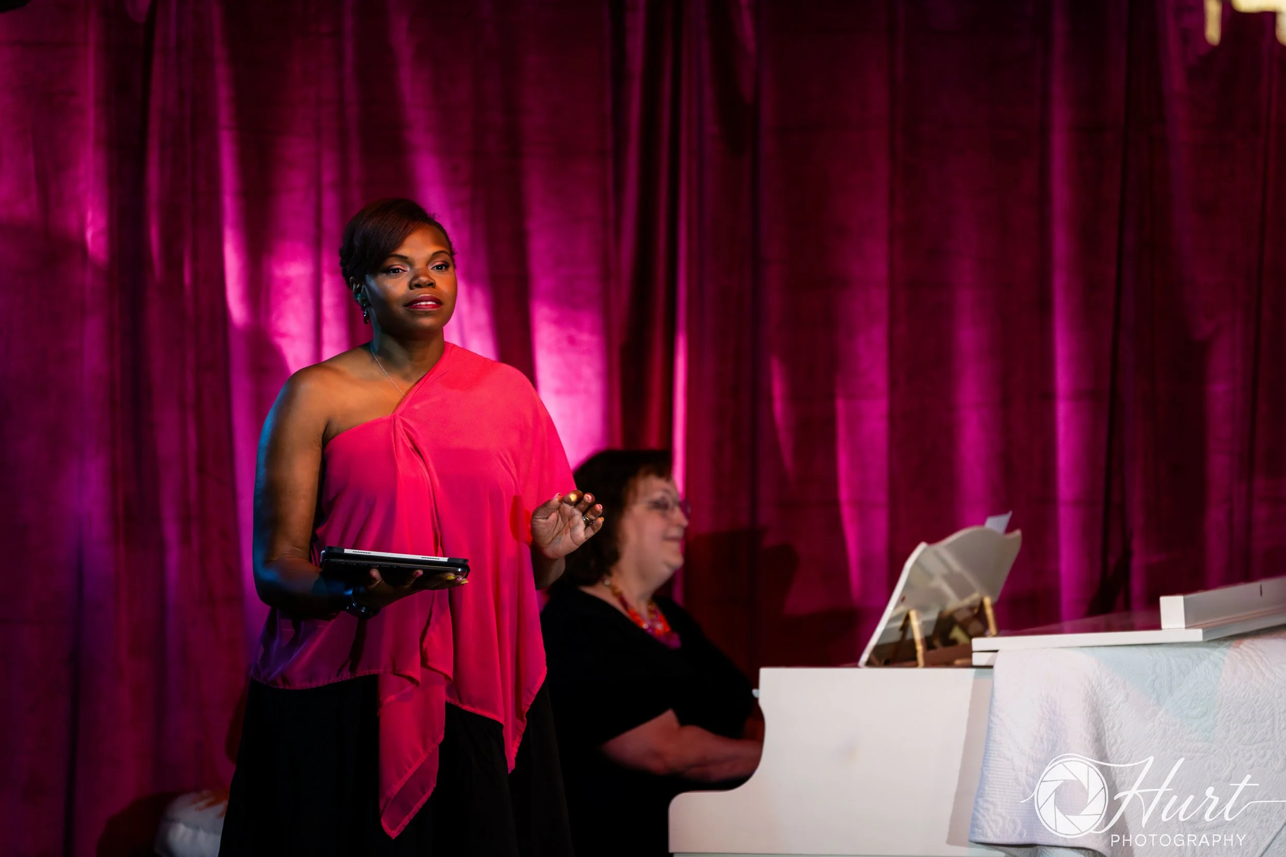 A woman in a pink dress is singing and holding a tablet standing in front of a magenta curtain, with another women sitting at a white piano in the background.