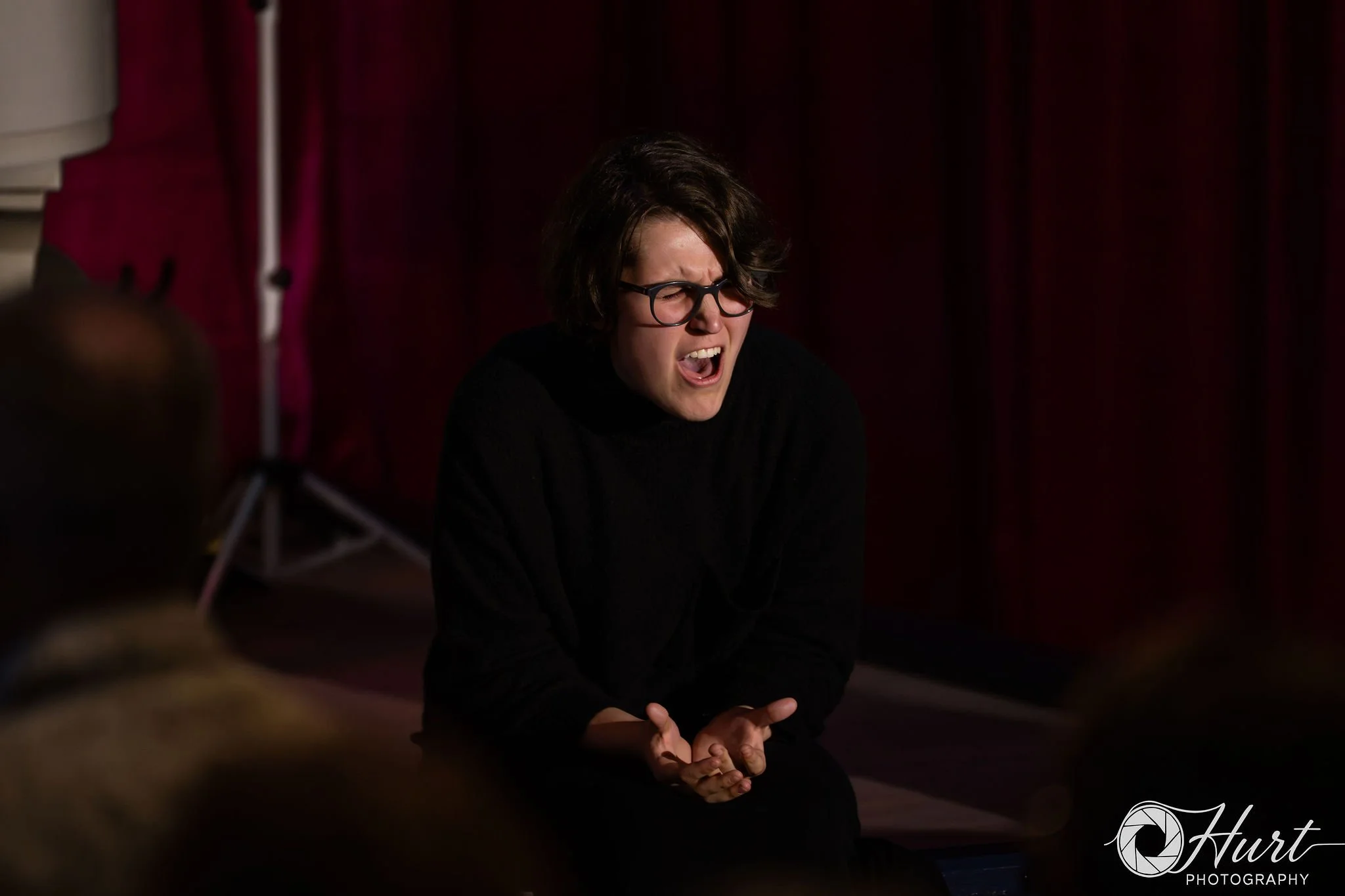 Woman with dark hair and glasses, sitting with her eyes closed and mouth open, expressing emotion, against a magenta curtain background.