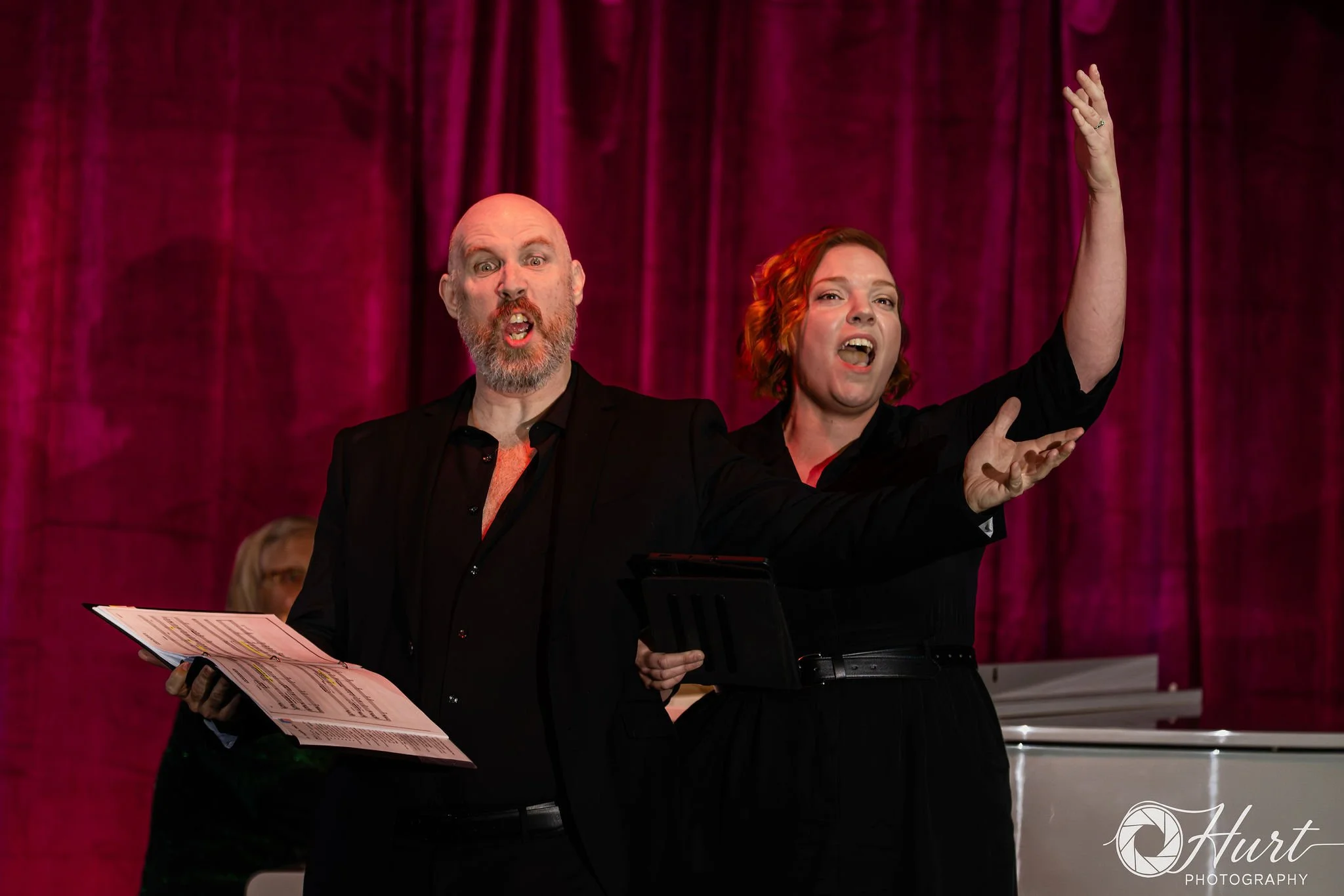 Two performers a man with a bald head and a beard, and a woman with curly red hair, are performing on stage in front of a magenta curtain. The man is holding sheet music and singing passionately, while the woman is raising her hand and also singing.
