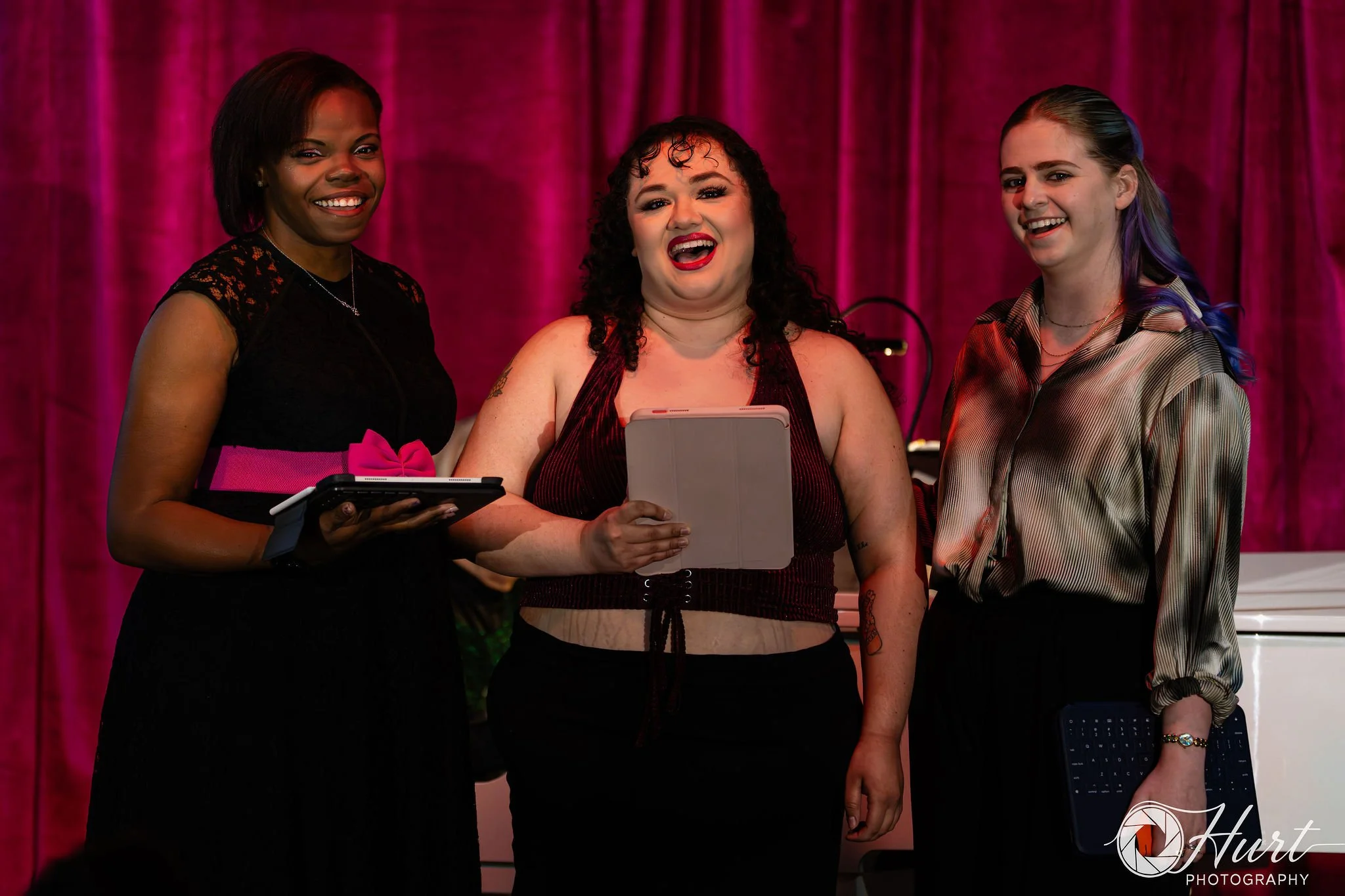 Three women standing in front of a magenta curtain singing. Left is an African American woman in a black dress with a magenta belt, middle is a Hispanic woman with curly hair in a black dress, right is a white woman wearing black and white.