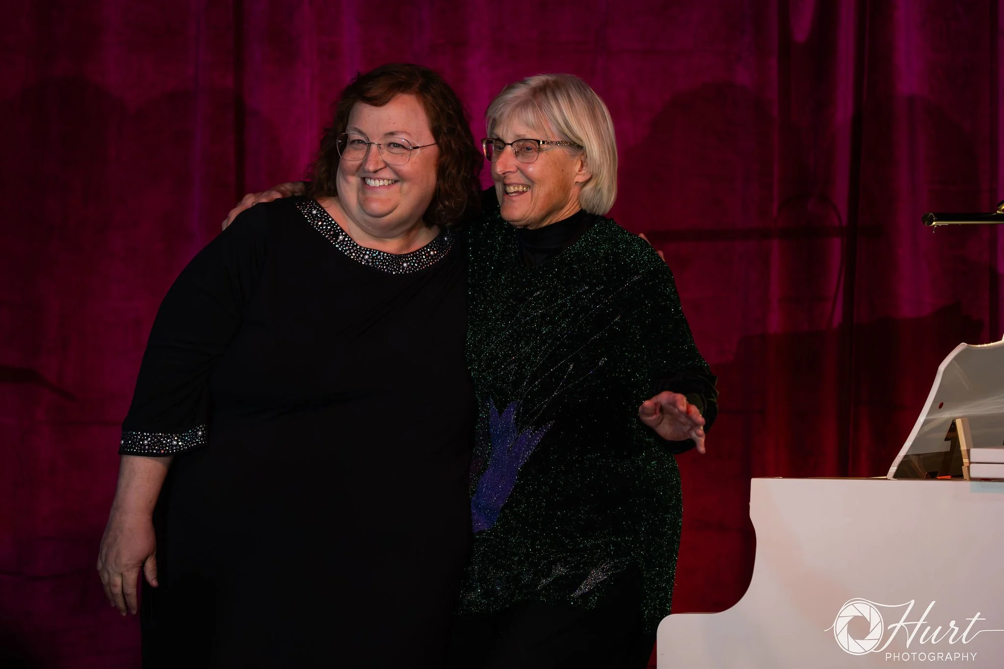 Two women stand at the white grand piano smiling and embracing each other on stage with a magenta curtain background.