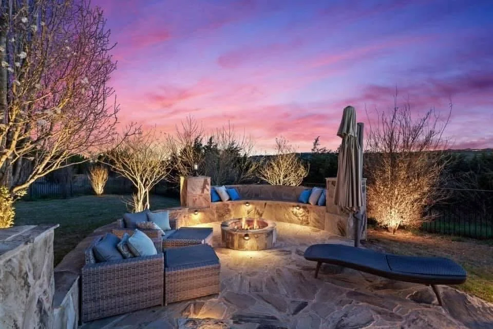 Outdoor patio area with wicker furniture, blue cushions, a fire pit, and a lounge chair, under a colorful sunset sky with pink and purple clouds, surrounded by trees.