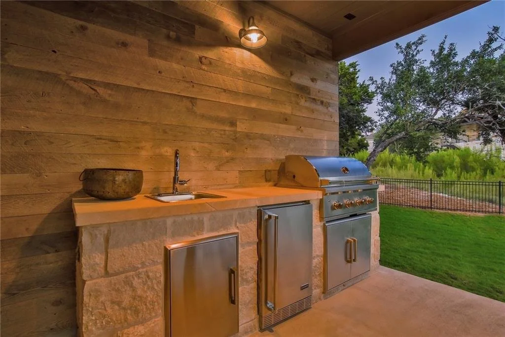 Outdoor kitchen area with a built-in grill, a small refrigerator, a stone counter with a sink, and a wooden wall backdrop, overlooking a grassy yard.