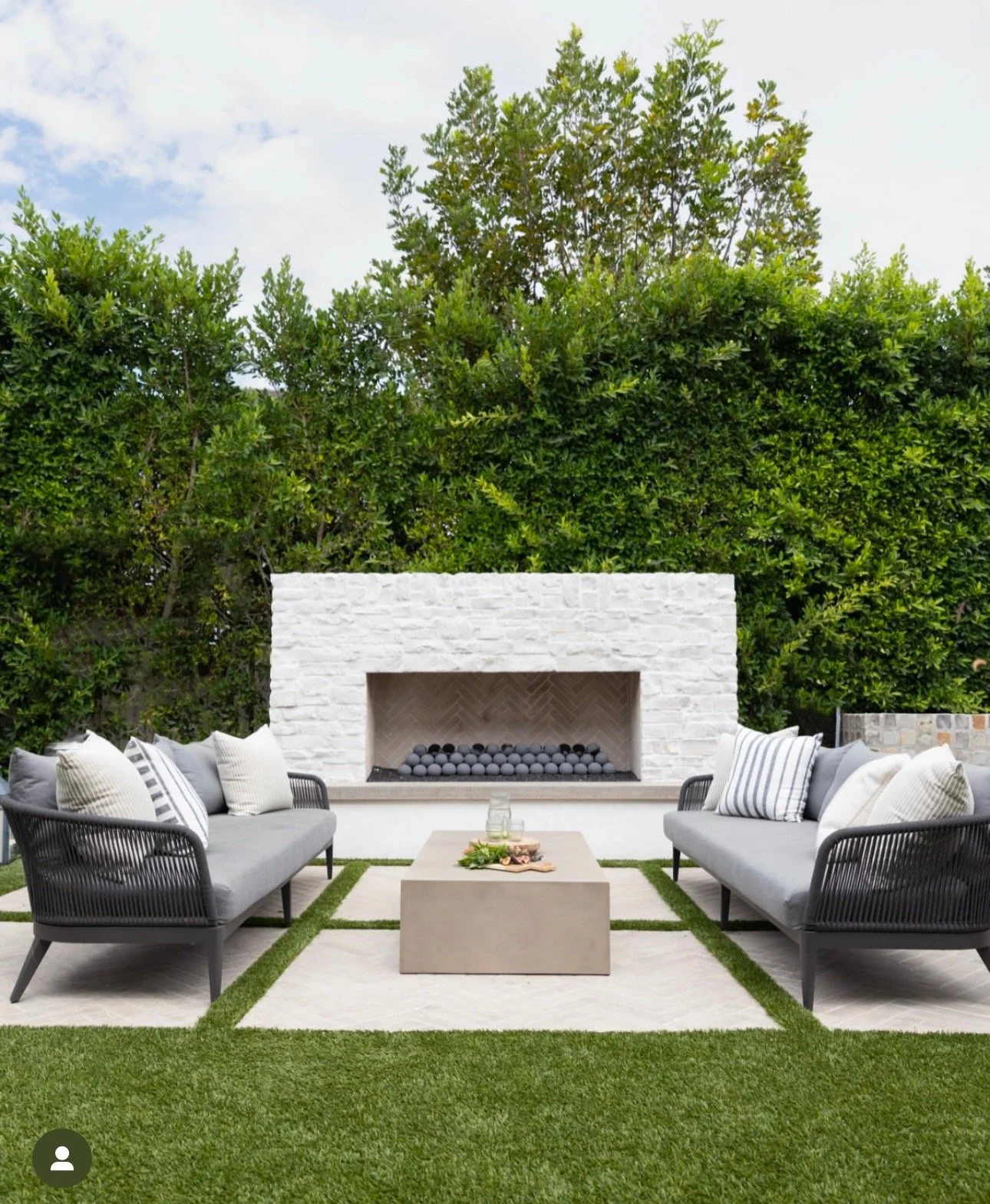 Outdoor patio with two gray couches, white and gray pillows, a concrete coffee table with a tray of drinks and snacks, a white brick fireplace with black decorative balls inside, and green hedges in the background.