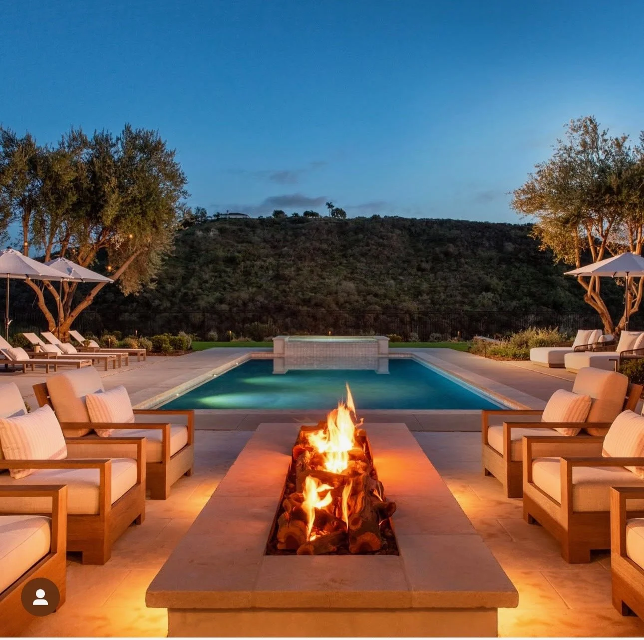 Luxury backyard with a swimming pool, outdoor seating, and a fire pit at sunset, surrounded by trees and hills.
