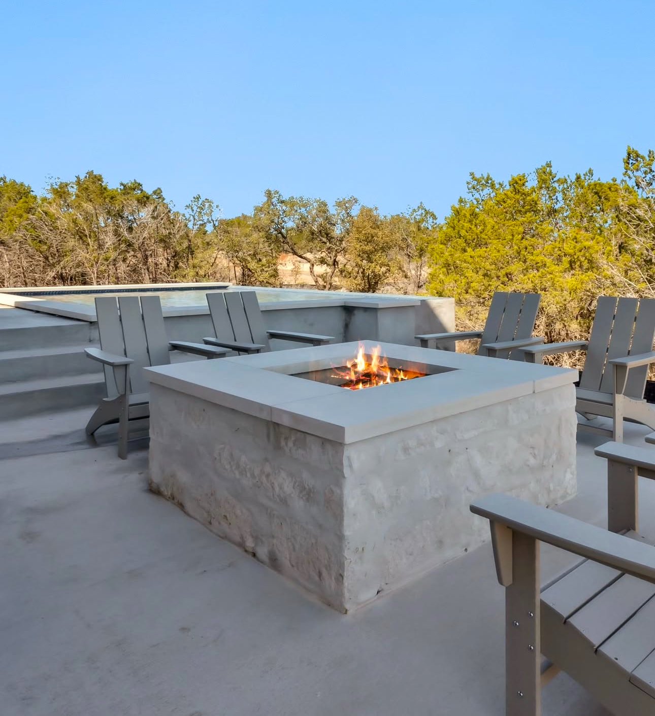 Outdoor patio with a stone fire pit surrounded by gray wooden chairs, overlooking trees under a clear blue sky.
