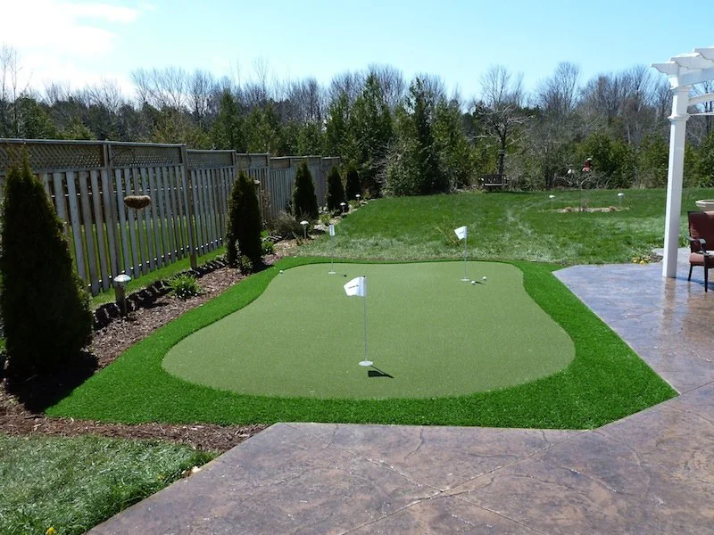 Backyard with a miniature golf putting green with three flags, surrounded by a fence and trees, next to a patio with outdoor furniture.