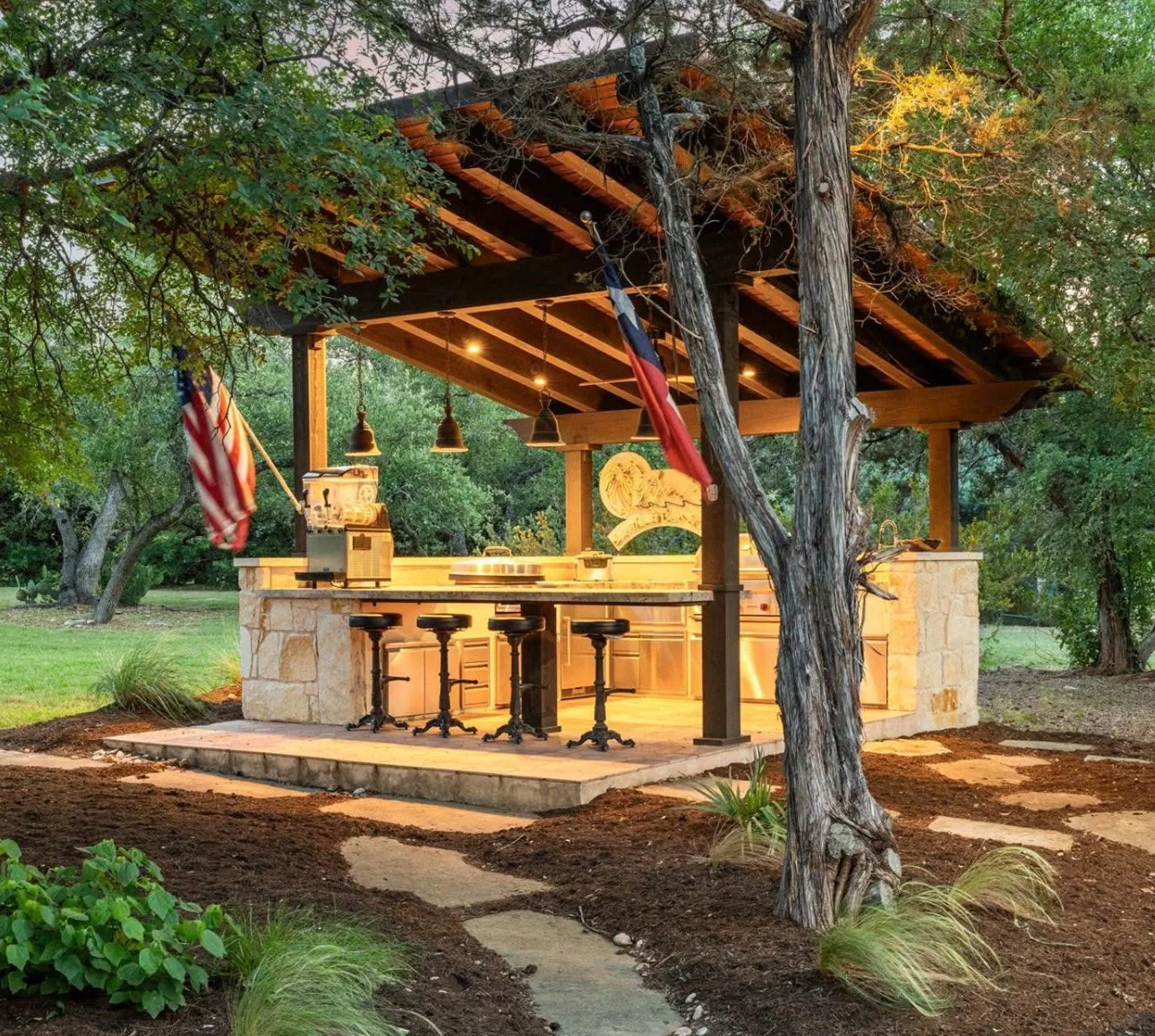 Outdoor kitchen with a stone counter, four black barstools, hanging pendant lights, and a roof with exposed wooden beams surrounded by trees and garden plants.