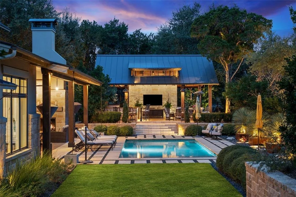 Luxury backyard at dusk featuring a swimming pool with illuminated water, surrounded by patio furniture, umbrellas, lush greenery, and a covered outdoor lounge area with a TV on a stone wall.