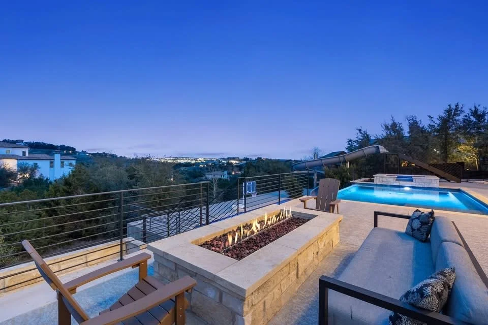 Outdoor rooftop patio at dusk with a swimming pool, fire feature, seating area, and city view in the distance.