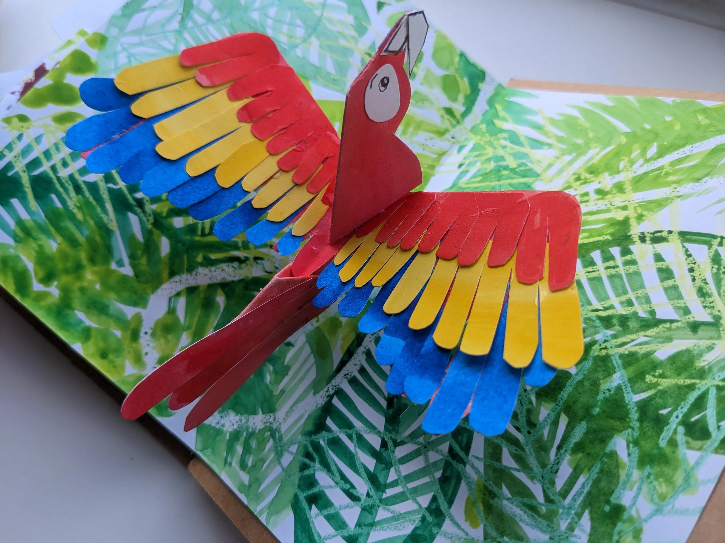 Colorful paper pop-up parrot with red, yellow, and blue feathers, on a green leafy background.
