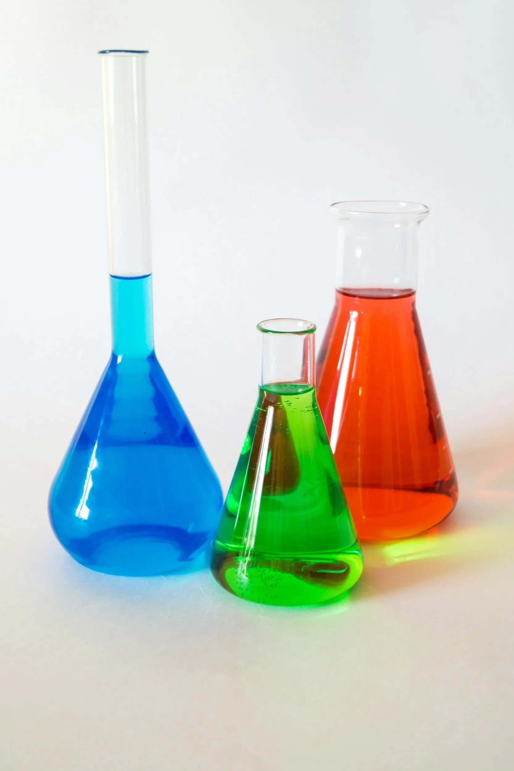 Four glass laboratory flasks containing brightly colored liquids: blue, green, red, and orange, arranged on a white surface with a plain white background.
