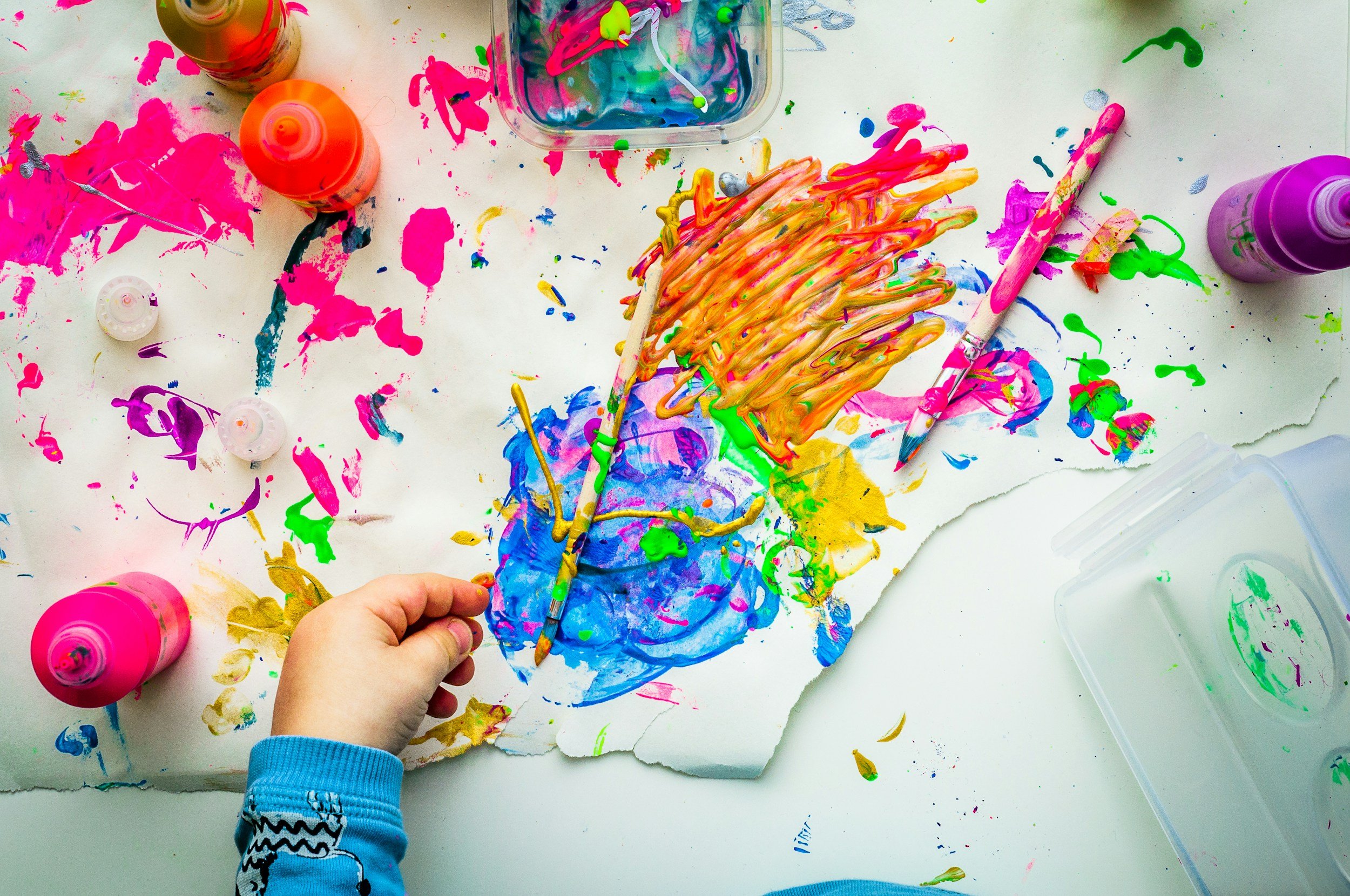 A child's hand holding a paintbrush with colorful paint on it, surrounded by spilled paint and art supplies on a white surface.