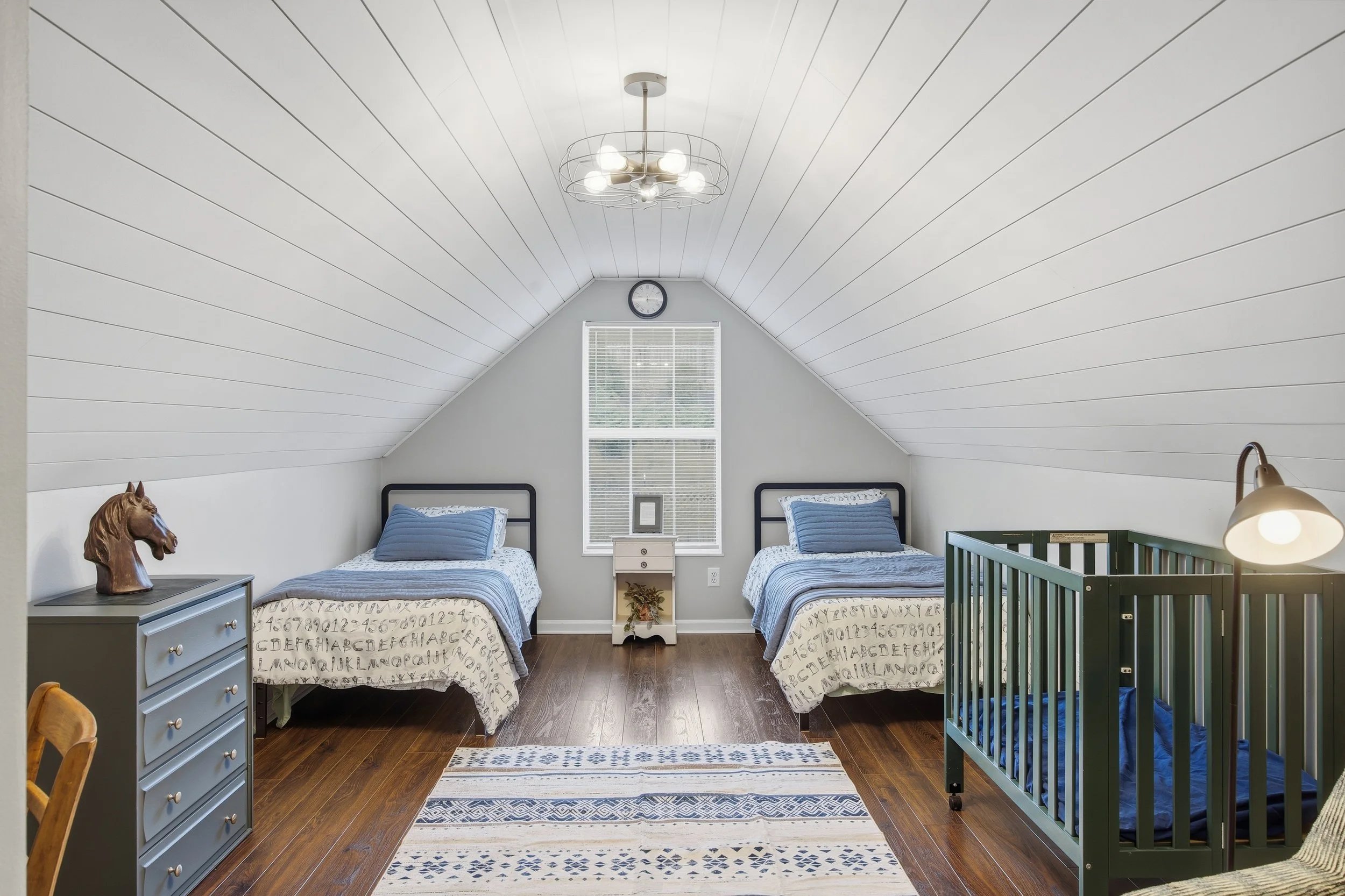 Bedroom with two twin beds with blue bedding, a small white nightstand between them, a window with blinds, a nursery crib on the right, and a gray dresser with a horse sculpture on top. The ceiling is white with a modern chandelier, and the floor is 