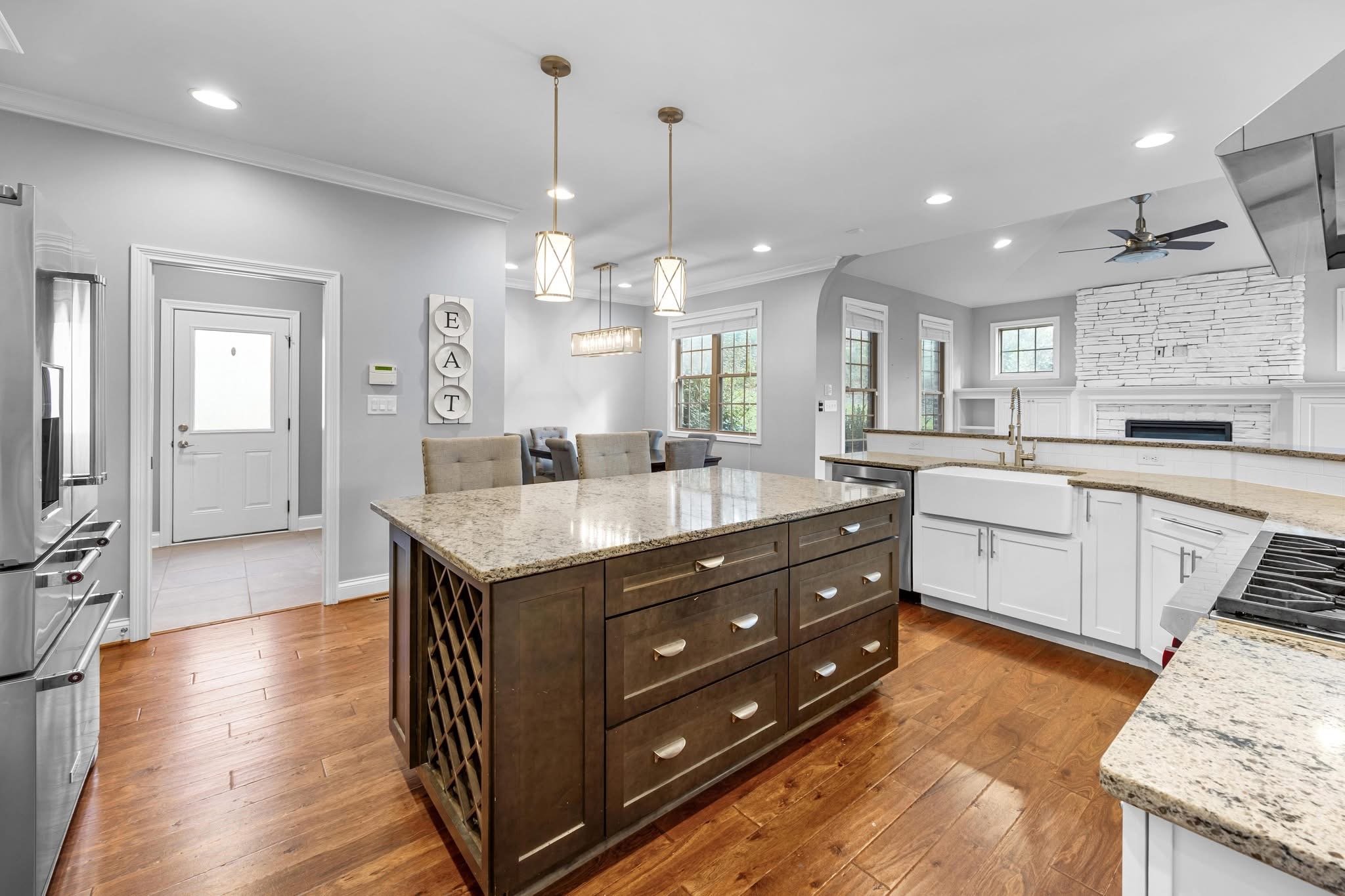 Modern kitchen with granite countertops, central island with drawers, white cabinets, stainless steel appliances, hardwood floors, large windows, and a white brick fireplace in the living room.