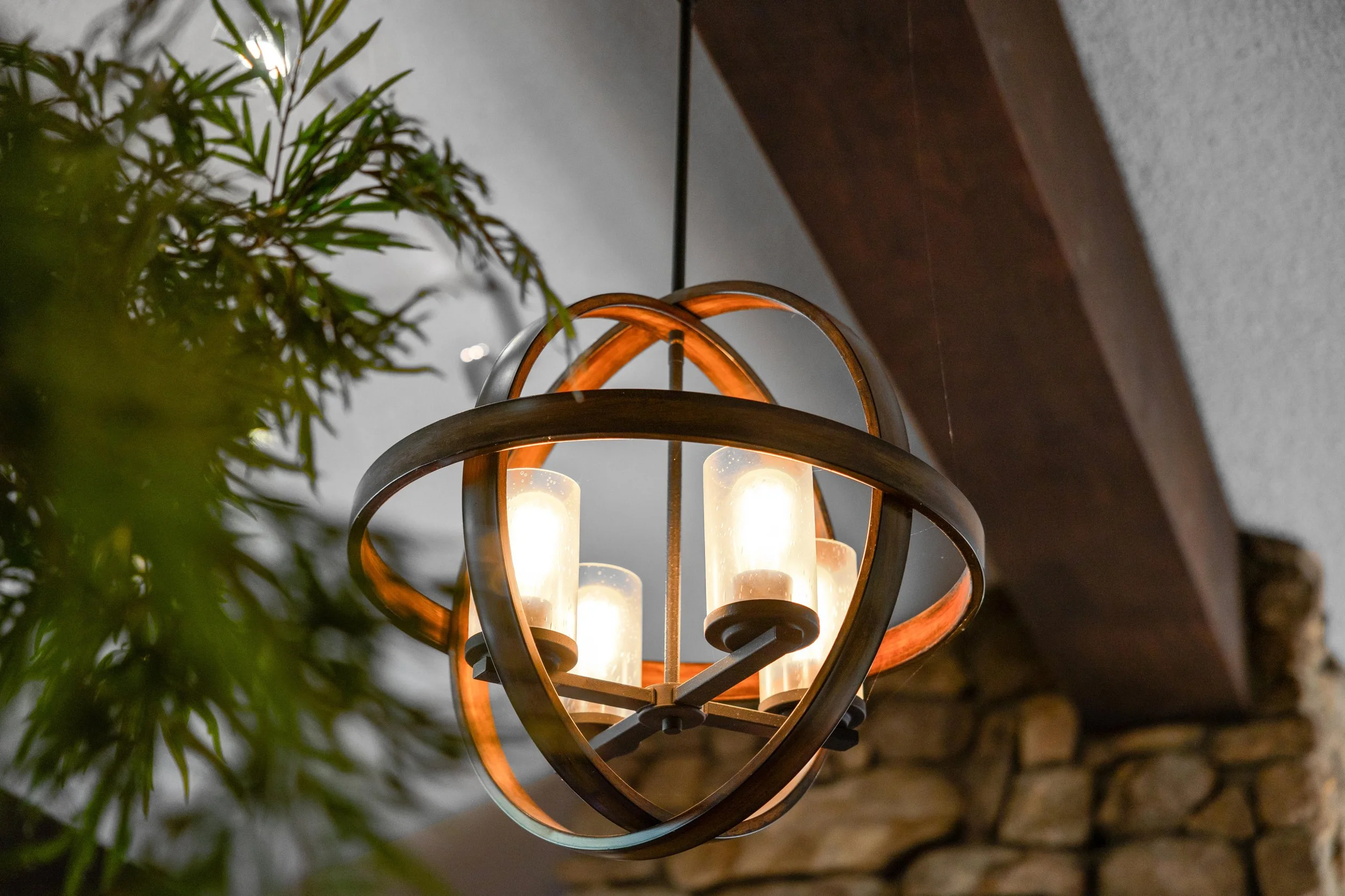 A modern chandelier with four candle-like light bulbs, surrounded by elliptical metal rings with a wooden finish, hanging from a ceiling with a gray wall and a stone wall in the background, and some green leaves partially visible in the foreground.
