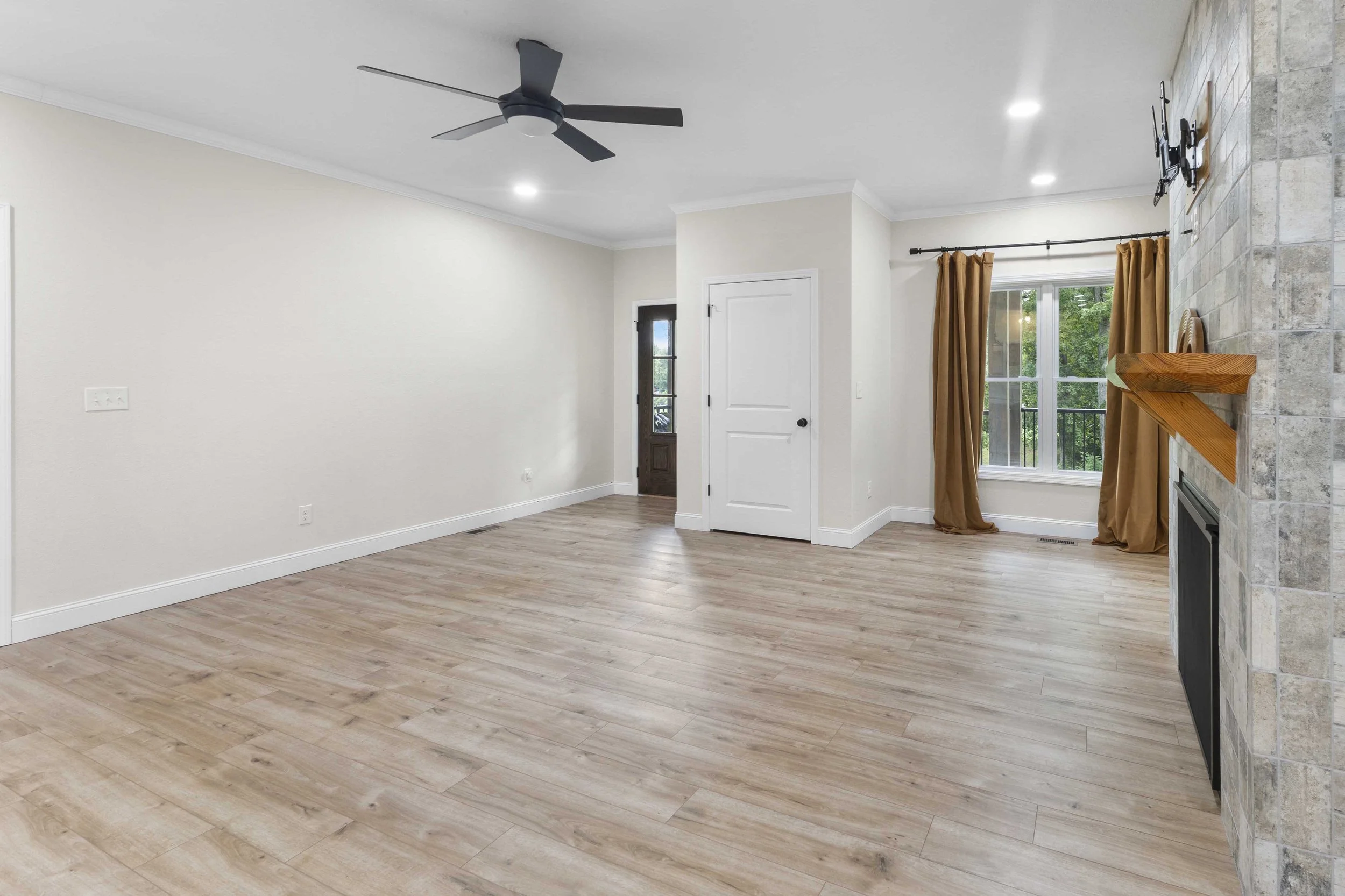 Empty living room with light wood flooring, white walls, a black ceiling fan, a window with tan curtains, a door, and a stone fireplace with a wooden mantel.