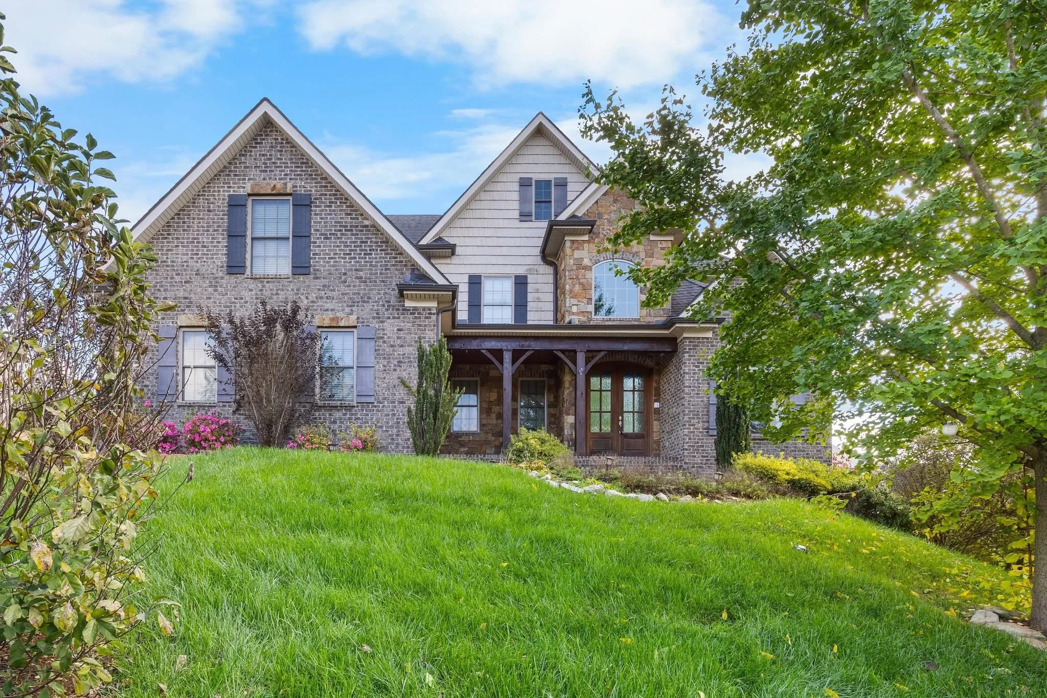 Front view of a large, two-story brick house with a front porch, surrounded by a green lawn, trees, and landscaped bushes under a partly cloudy sky.