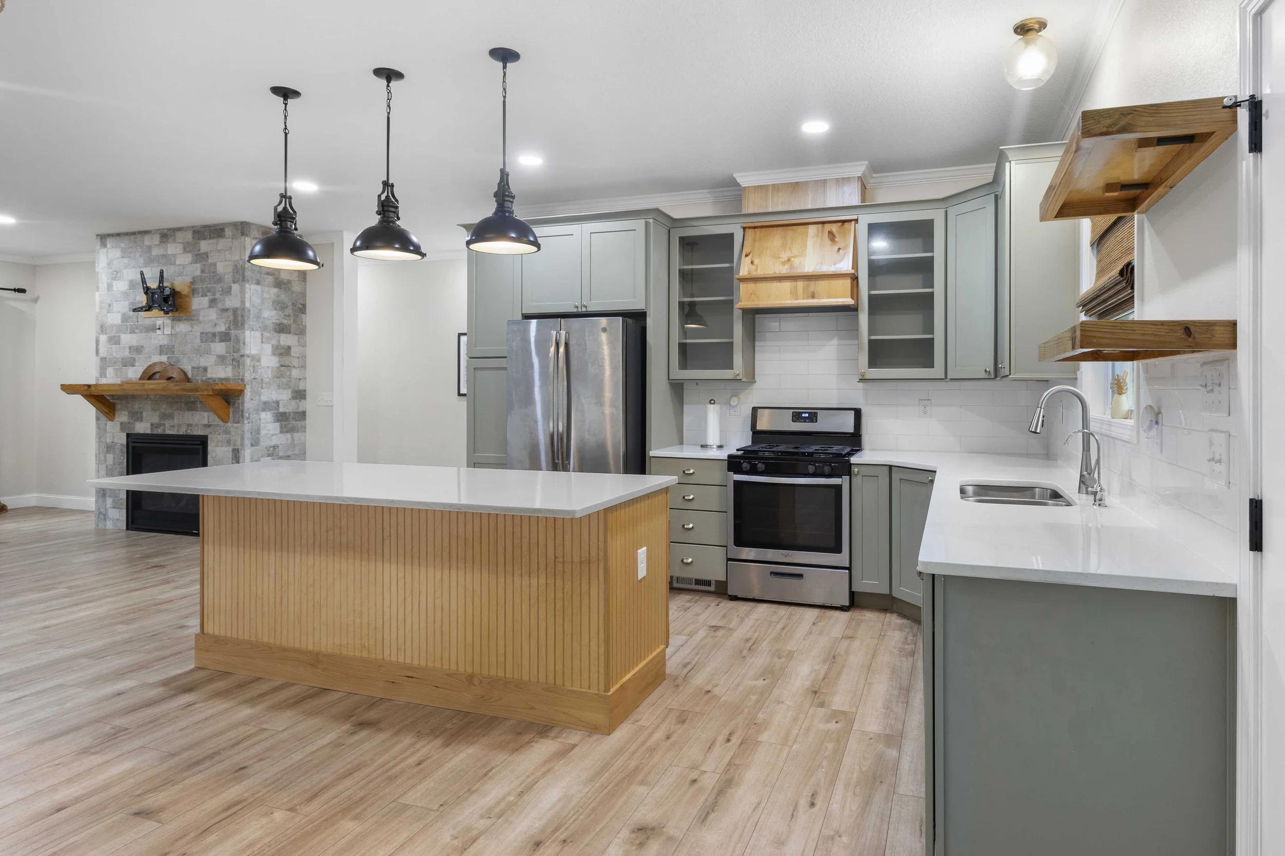 Kitchen with green cabinets, stainless steel refrigerator and stove, white countertops, wooden kitchen island, hardwood floors, open shelves, and a stone fireplace with a wooden mantle.