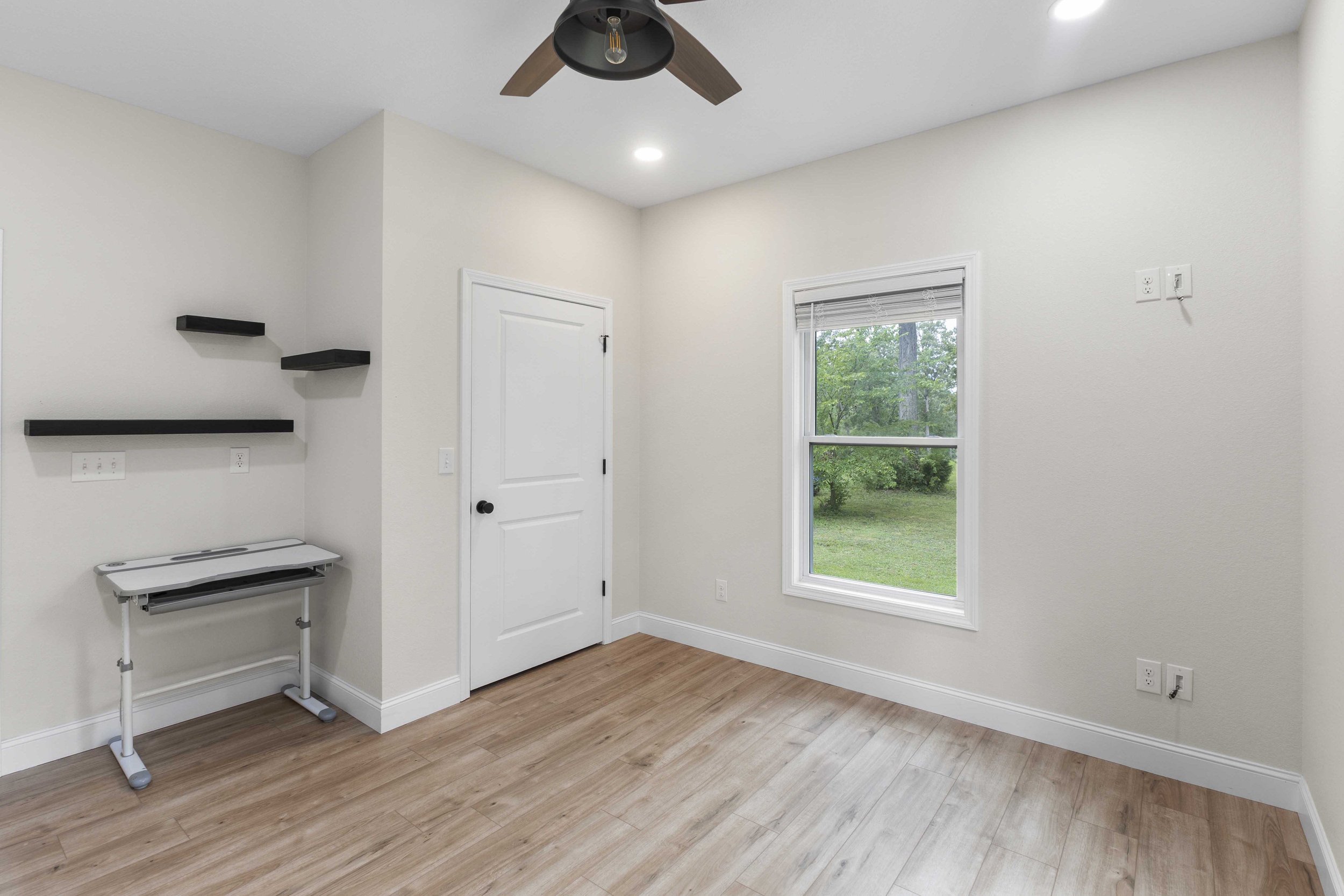 Empty room with a window showing greenery outside, a ceiling fan, and shelves on the wall.