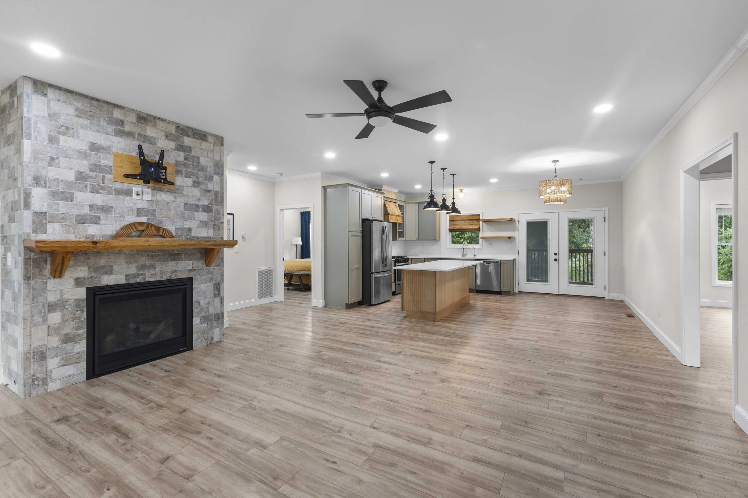 Open living area with kitchen, fireplace with wooden mantel, pendant lights, ceiling fan, and French doors leading outside.
