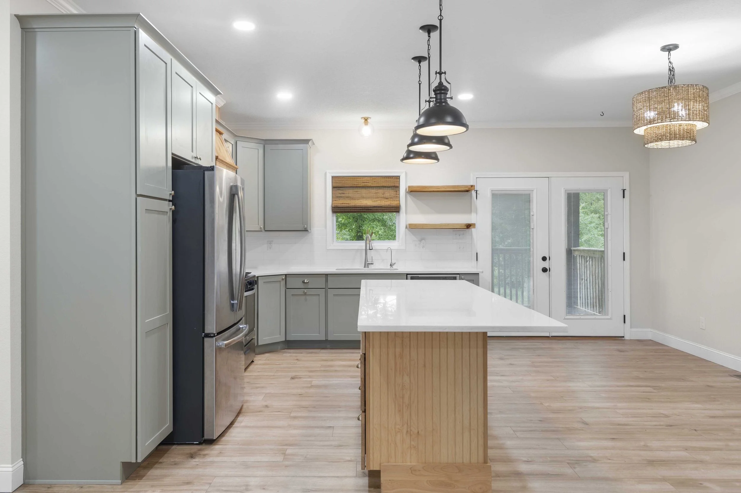 Modern kitchen with light gray cabinets, a stainless steel refrigerator, white countertop, island with wooden base, and a small window with a roller shade, open shelving, and a glass door leading outside.