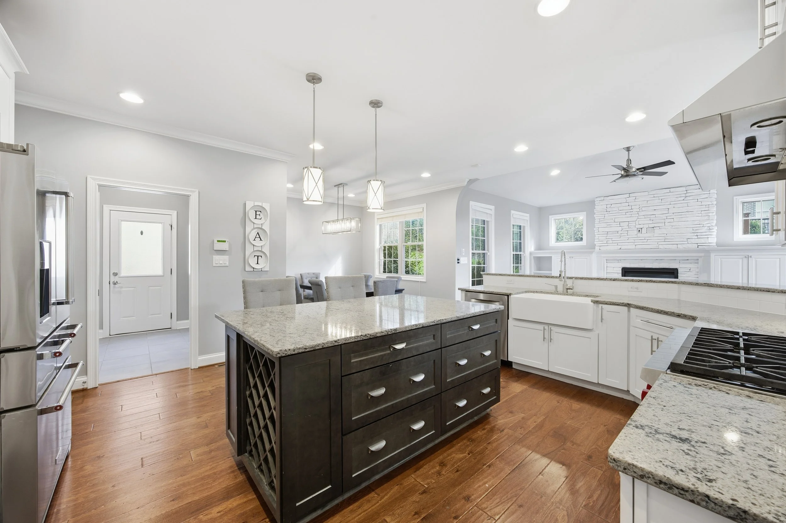 Modern kitchen with granite countertops, white cabinetry, and a central island with dark wood drawers. The space features hardwood floors, stainless steel appliances, and a bright, open layout with large windows. A dining area with a table and chairs
