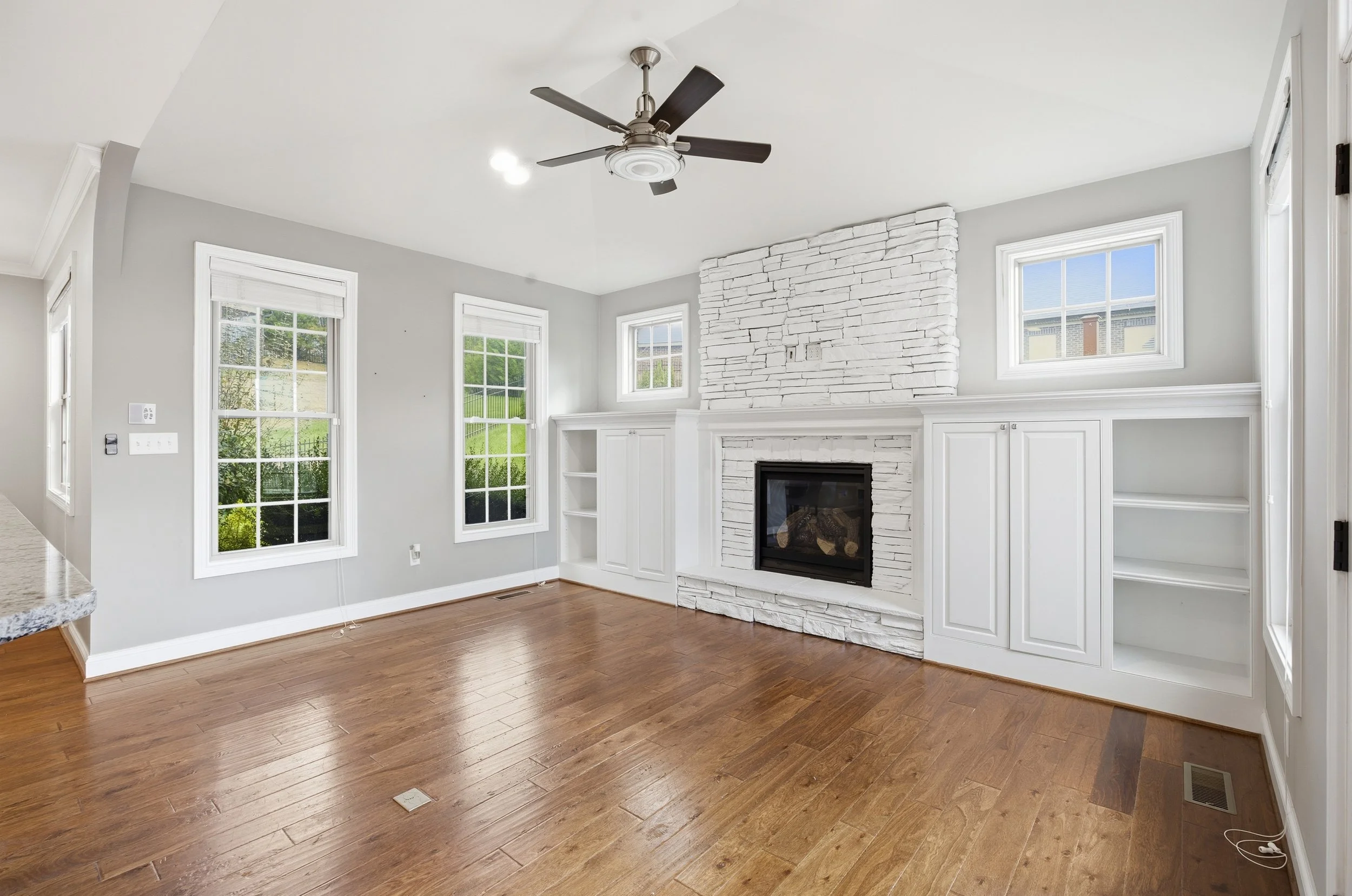 Empty living room with hardwood floors, white walls, three large windows, a white stone fireplace, cabinets on either side, and a ceiling fan.