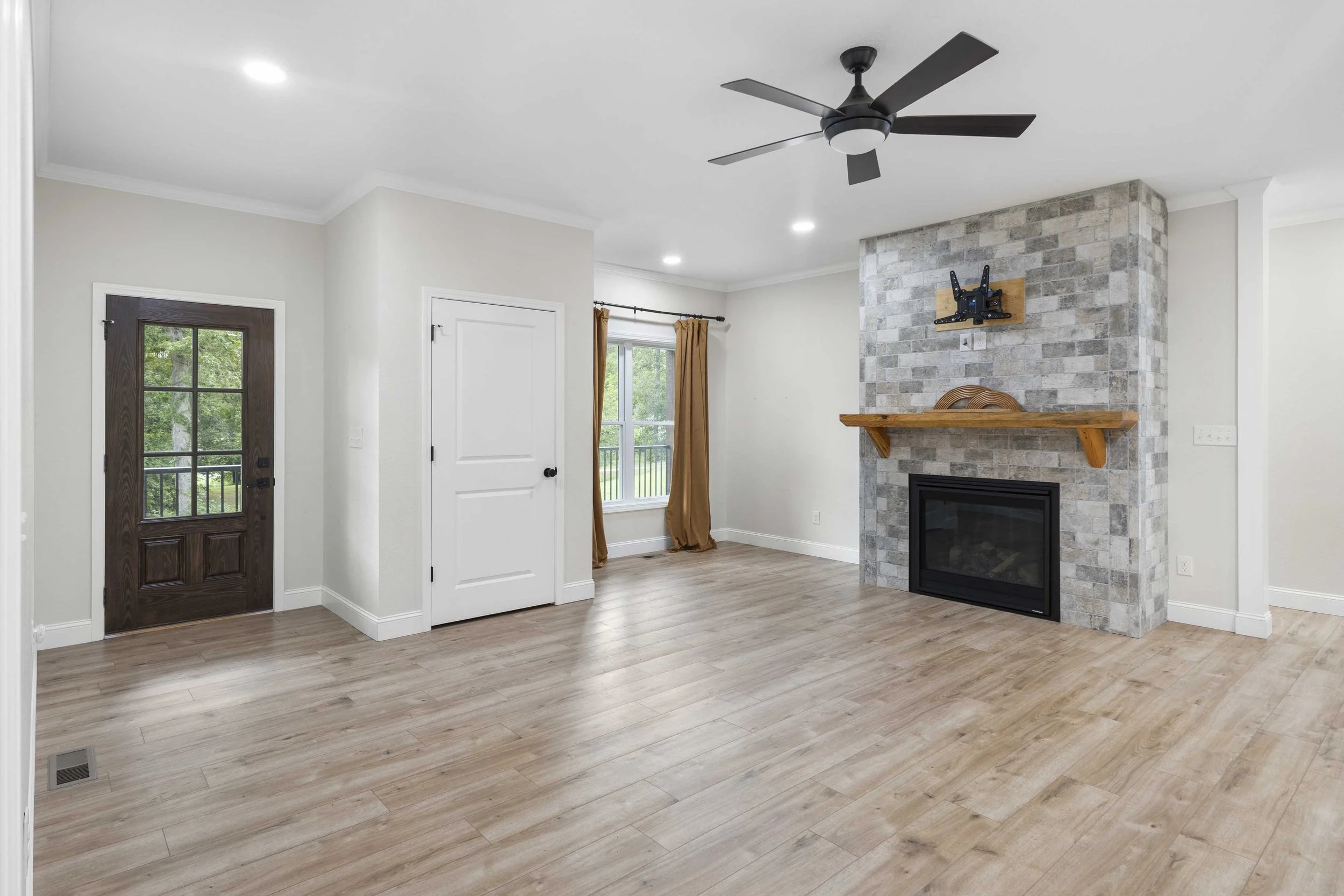 Empty living room with wood flooring, a fireplace with a stone surround and wooden mantel, ceiling fan, windows with gold curtains, a wooden door with glass pane, and a white door in the corner.