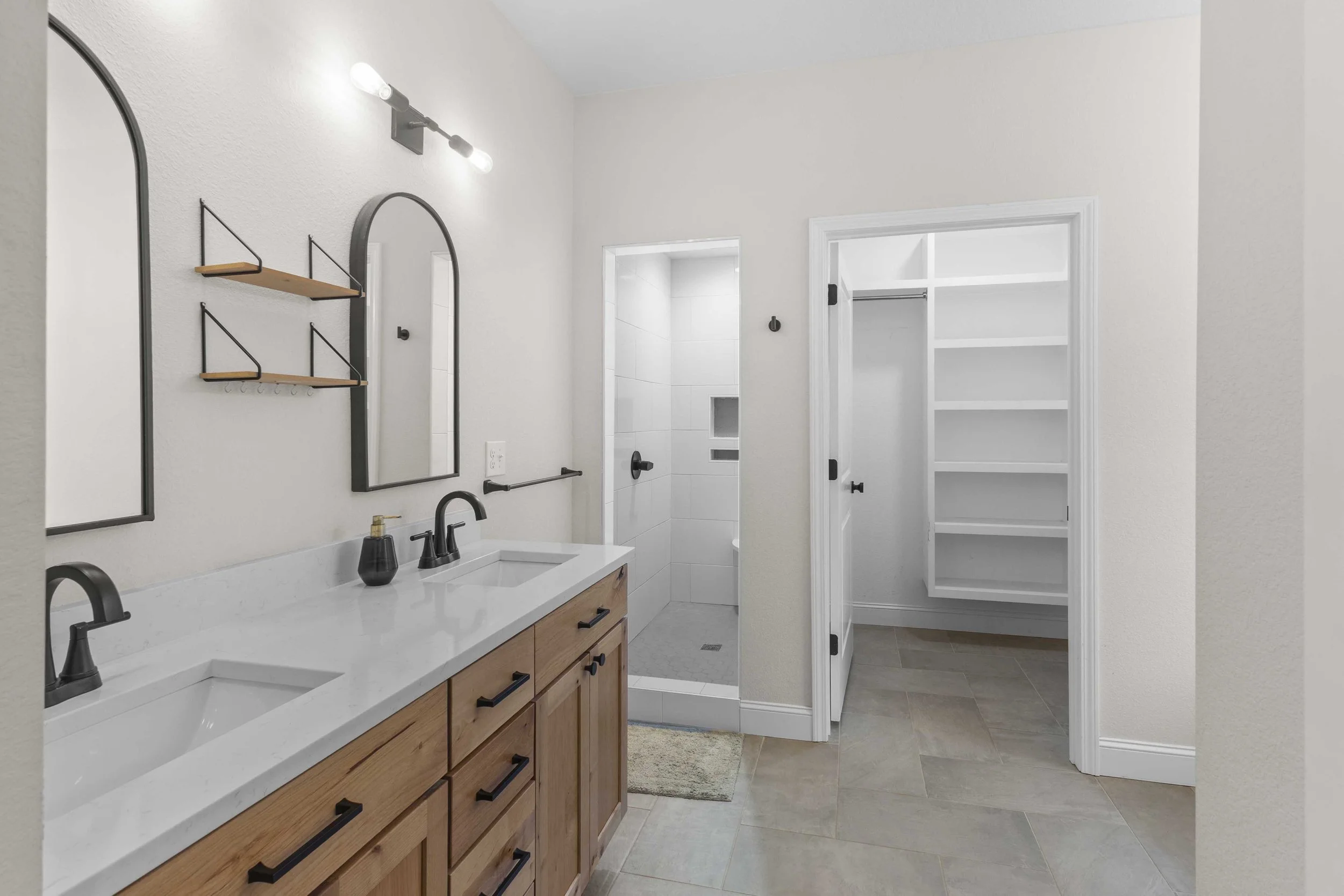Modern bathroom with double vanity, two sinks, wooden cabinets, black fixtures, mirrors, shelves, shower area, and white shelving closet.