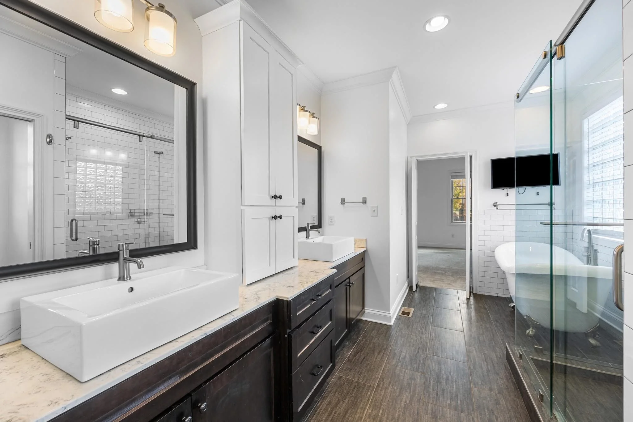 Modern bathroom with double vanity sinks, large mirror, white cabinetry, and a separate glass-enclosed shower and soaking tub, with a wall-mounted TV.