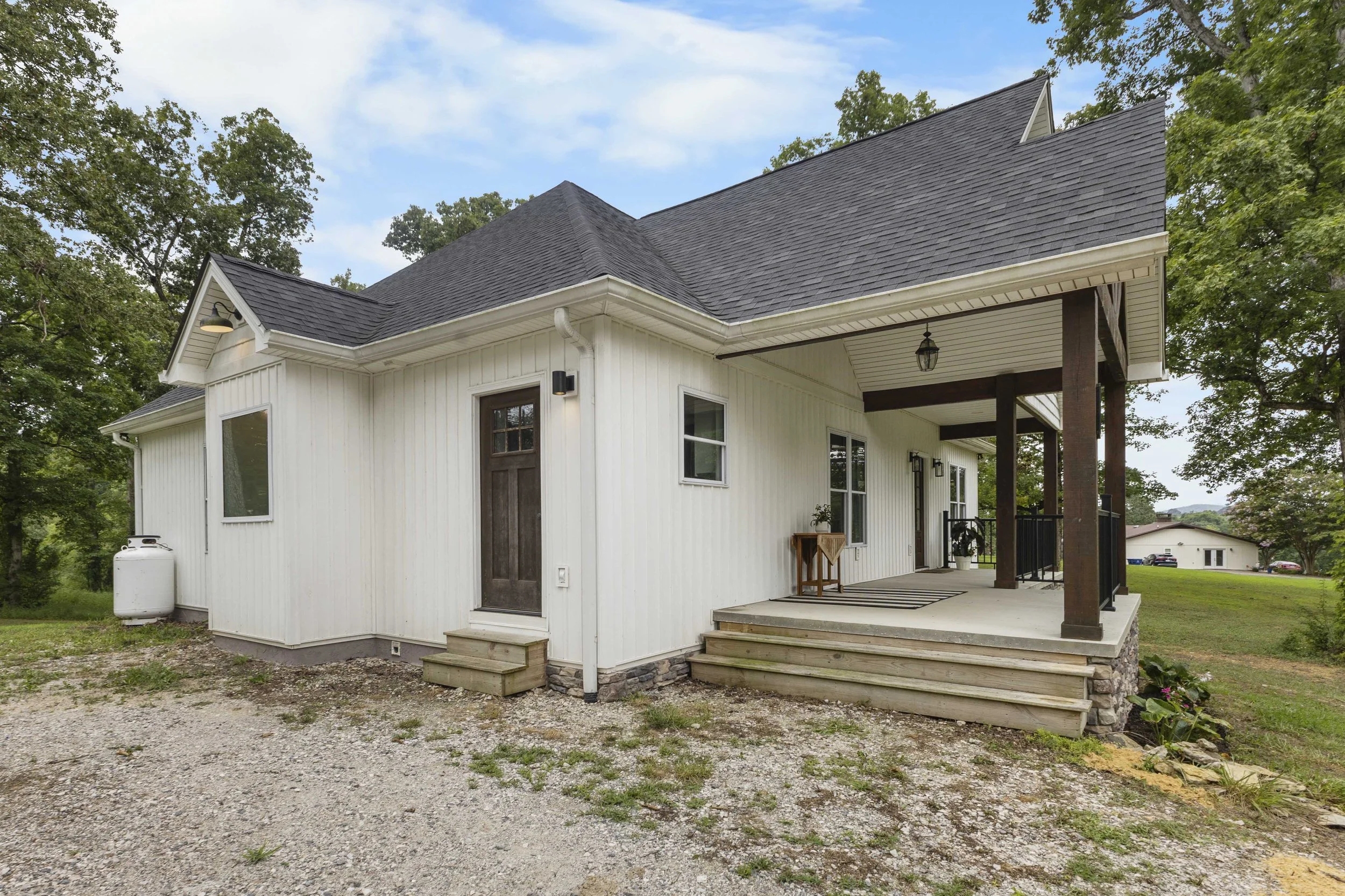 White house with a covered porch, dark brown front door, black railing, and steps leading up to the porch, surrounded by trees, under a partly cloudy sky.