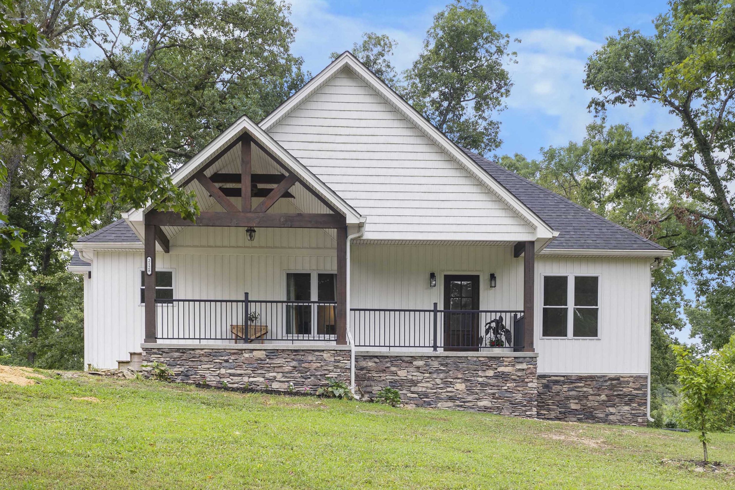 White house with a wooden stone foundation, covered porch, and dark roof, surrounded by green trees and grass.