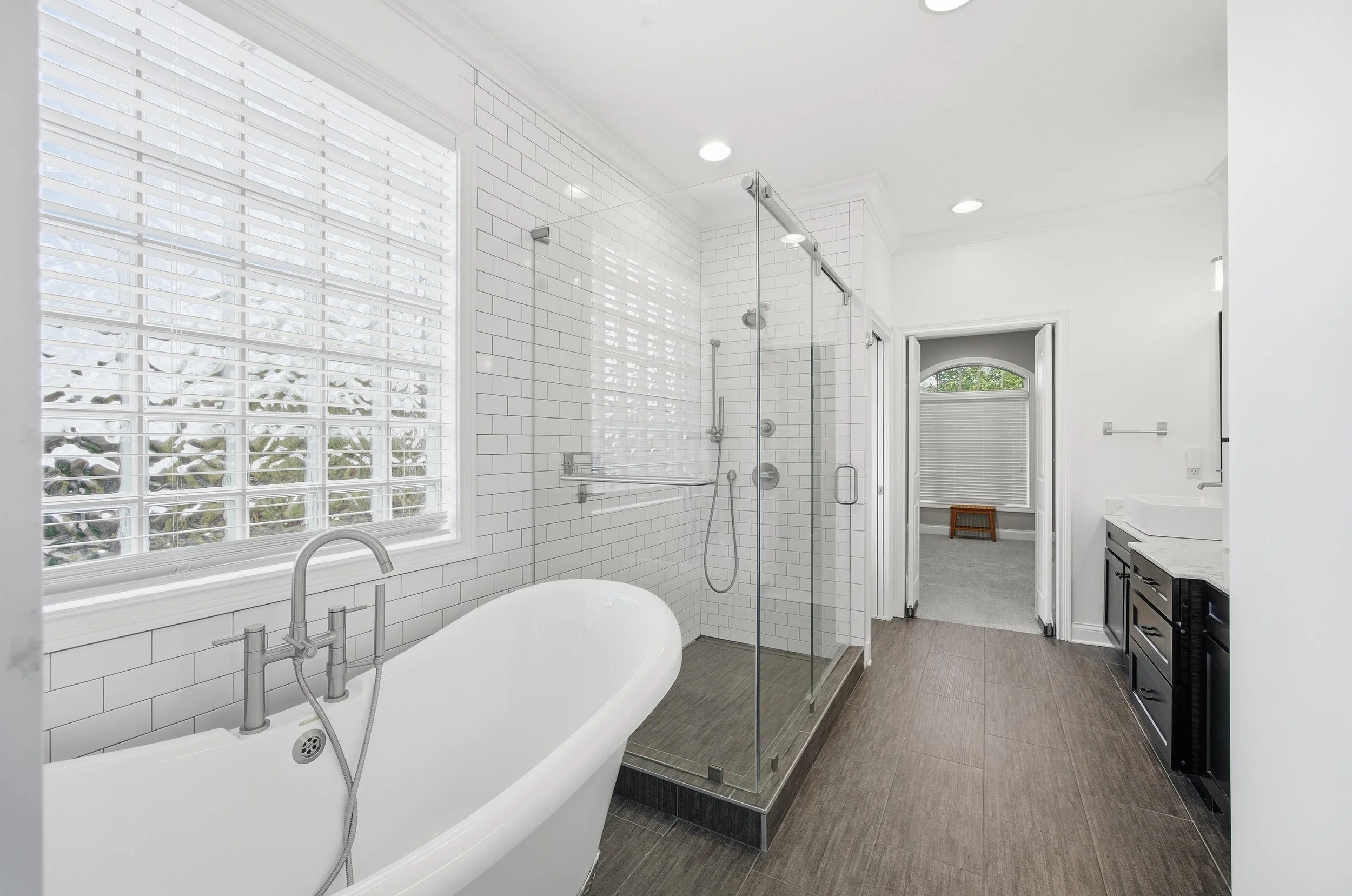 A modern bathroom featuring a white freestanding bathtub, a glass-enclosed walk-in shower with white subway tiles, a window with white plantation shutters, and a dark wood vanity with a marble countertop.