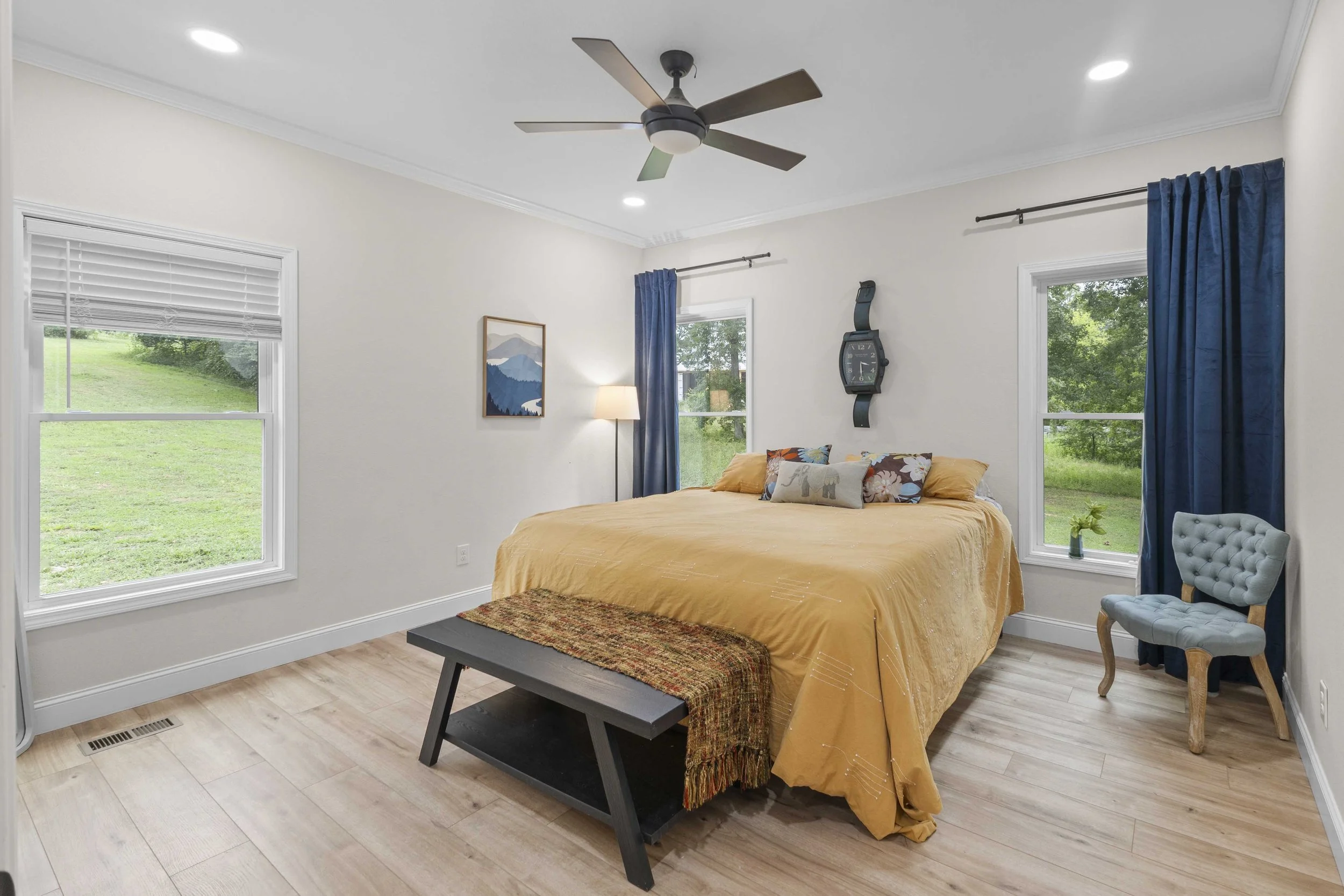 Bedroom with a large bed covered with a yellow bedspread, decorative pillows, a small bench at the foot, blue curtains, two windows showing a green landscape, a wall clock, a framed picture, a floor lamp, and a light blue chair.