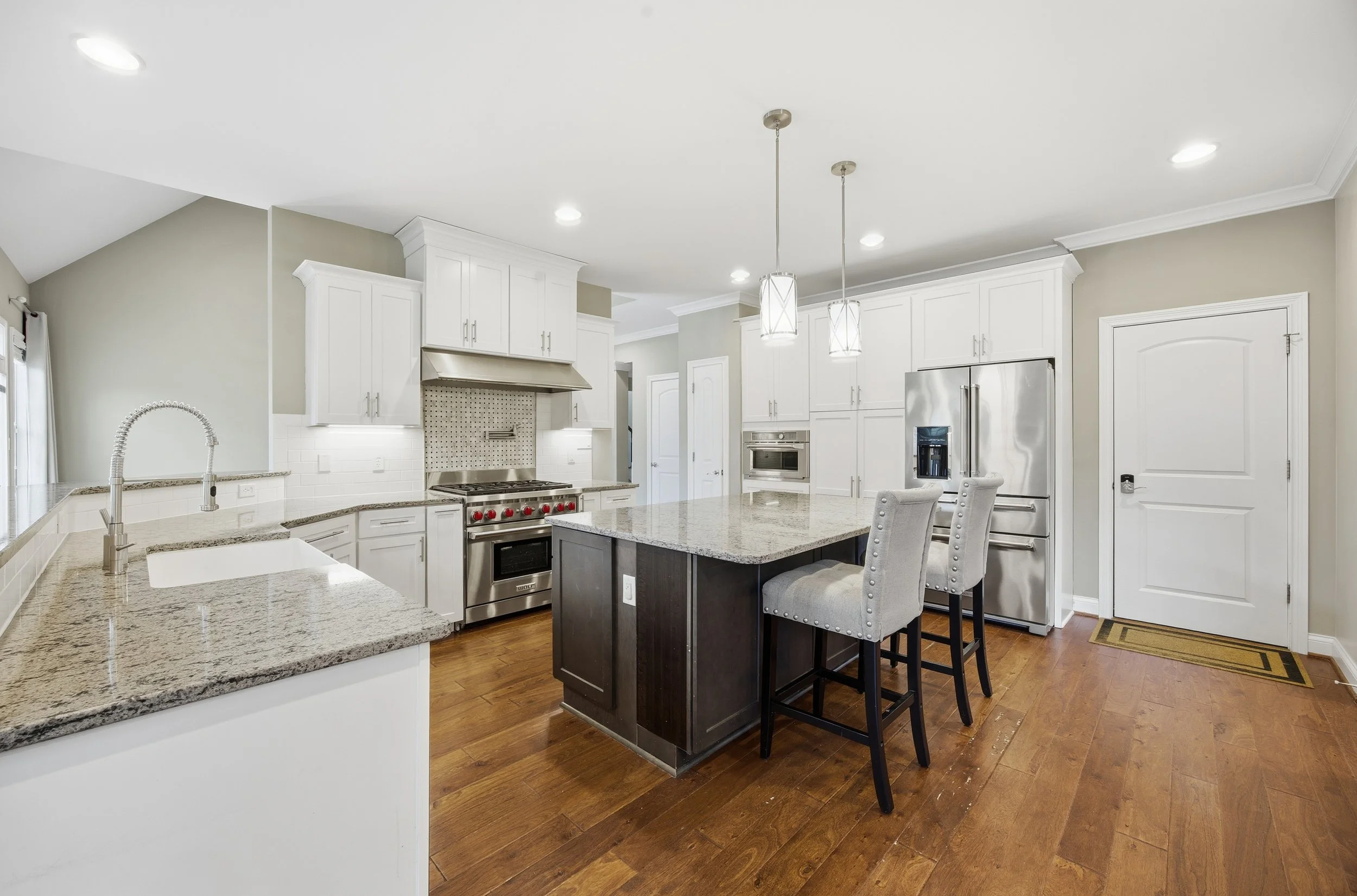 Modern kitchen with white cabinetry, stainless steel appliances, granite countertops, and a kitchen island with seating. Wooden flooring and pendant lights above the island.