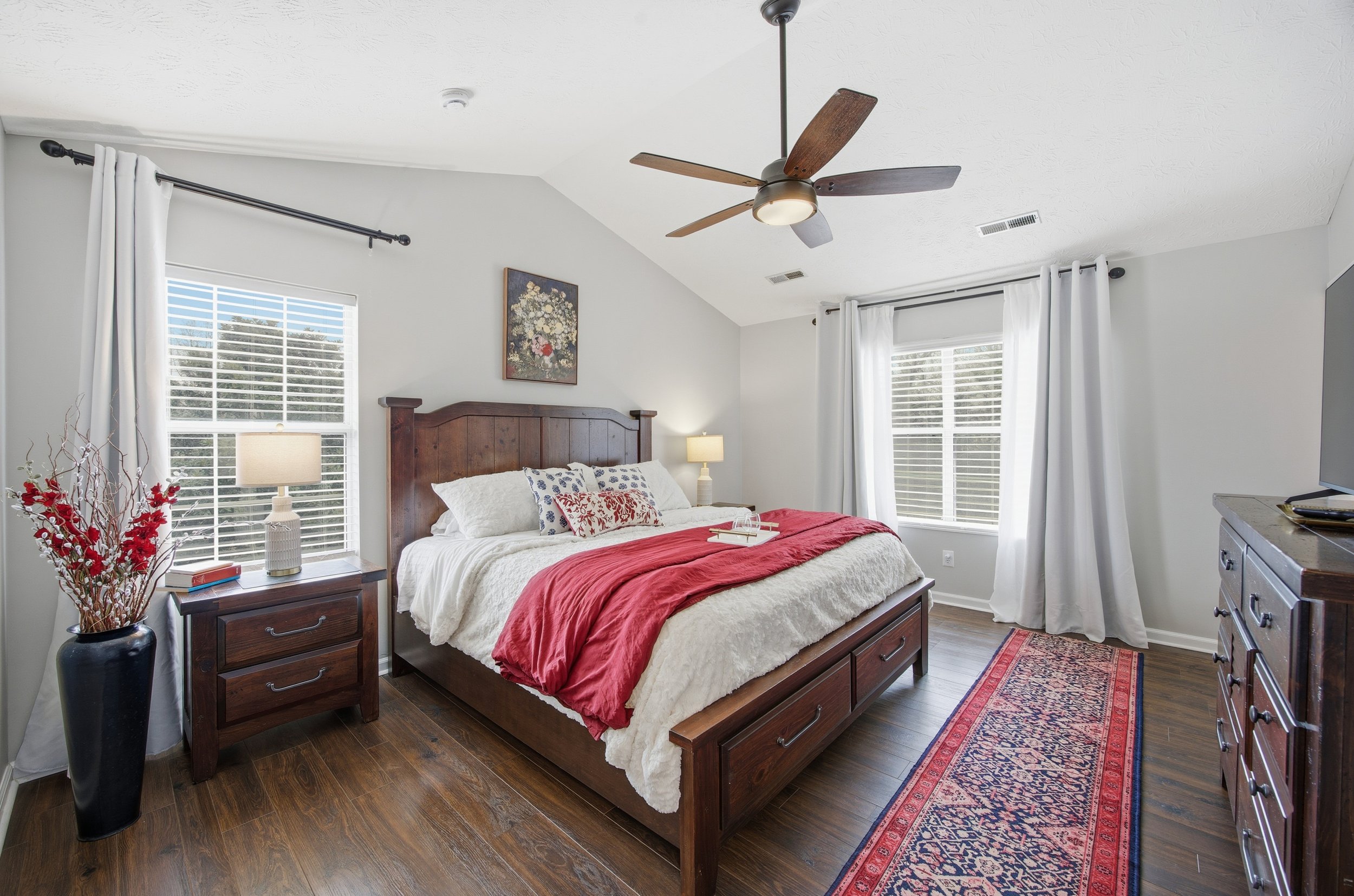 A bedroom with a wooden bed frame, white bedding, multiple pillows, red blanket, two nightstands with lamps, a ceiling fan, windows with white curtains, a gray dresser, and a decorative rug.
