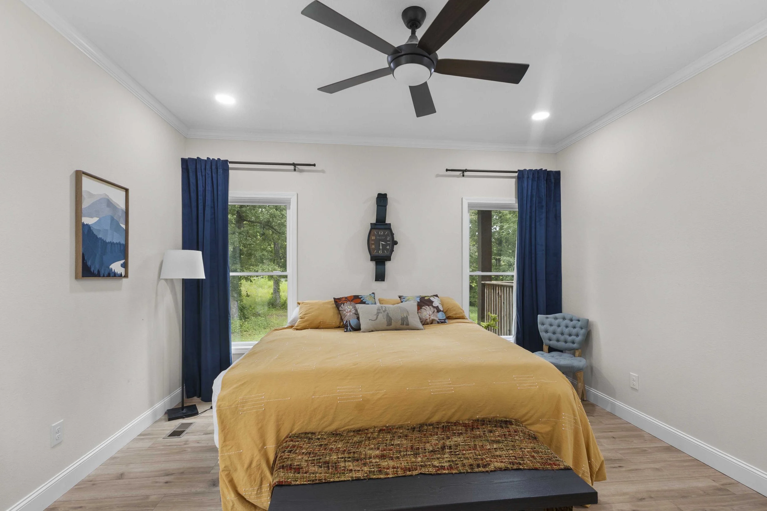 A bedroom with a large bed covered in a yellow bedspread and decorative pillows, two windows with blue curtains, a ceiling fan, a wall clock, a small blue tufted chair, and a floor lamp.