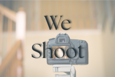 A camera mounted on a tripod or stabilizer in front of a blurred background with the text 'We shoot' on the wall.