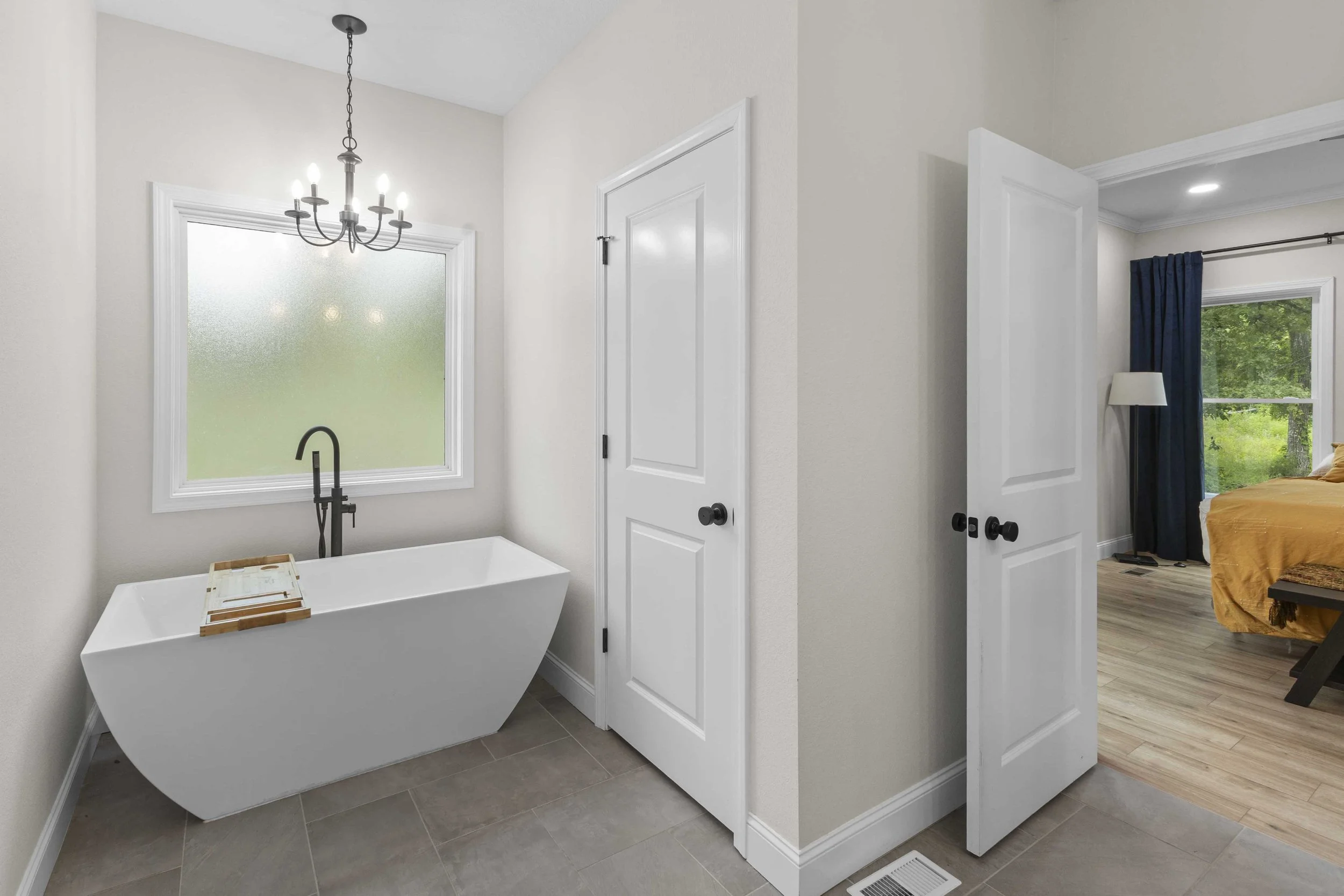 Bathroom with a freestanding bathtub under a frosted window, chandelier overhead, and a door leading to a bedroom with a yellow bedspread and window with dark curtains.