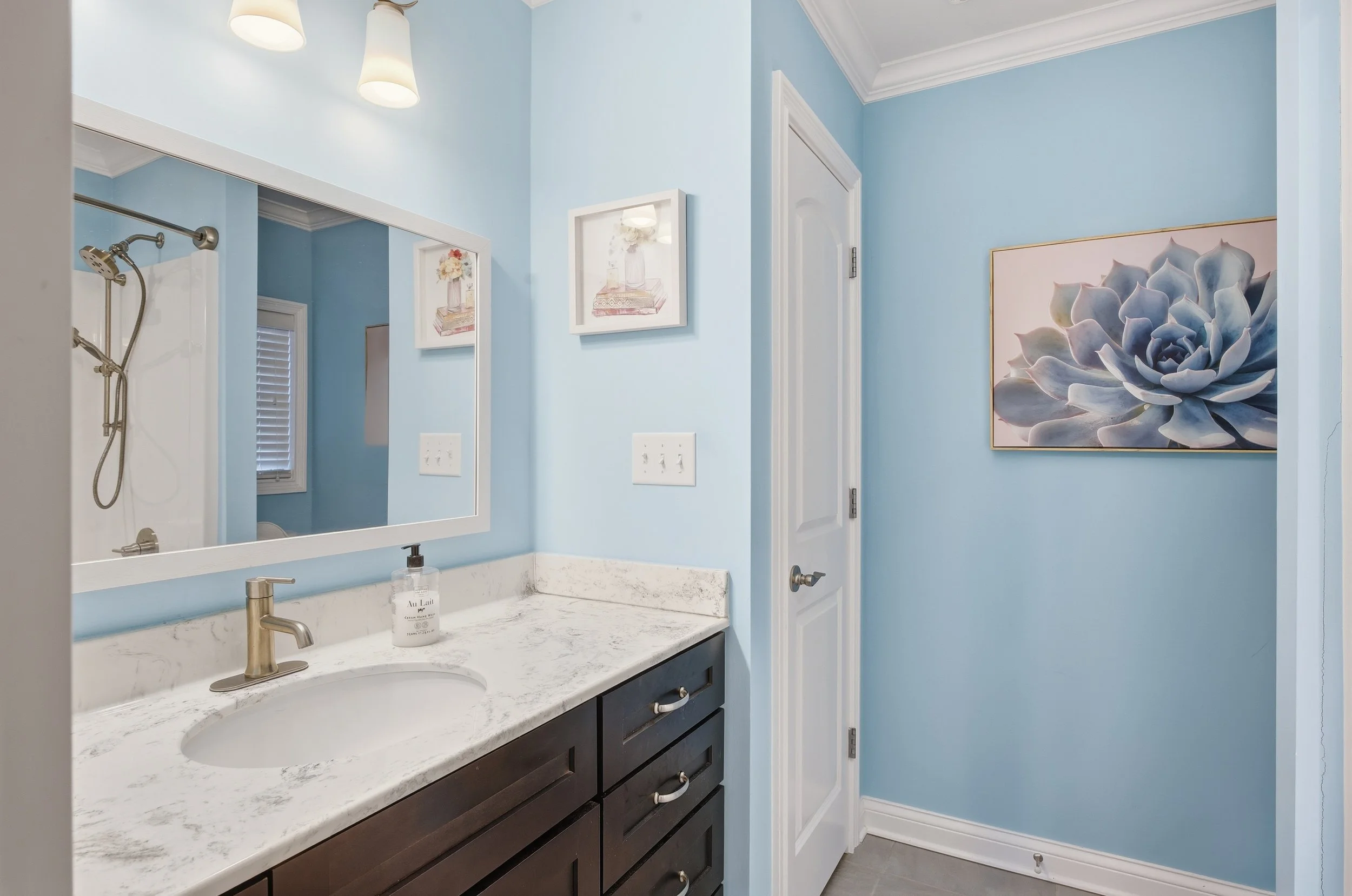 A bathroom with light blue walls, a large mirror above a marble countertop with a sink, dark wood cabinets, a hand soap dispenser, and artwork including a large flower painting on the wall.