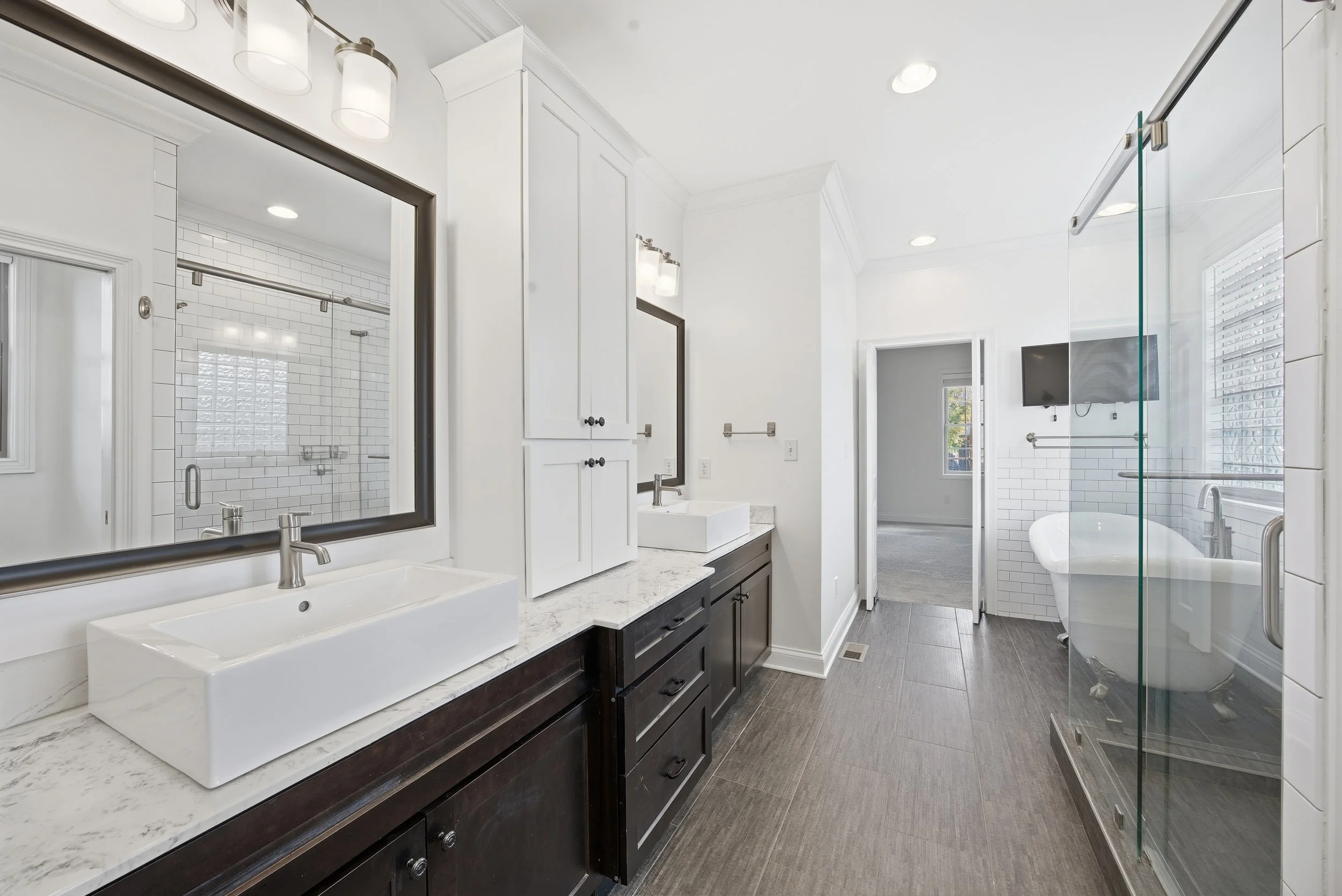 Modern bathroom with white walls, dark wood cabinets, white vessel sinks, large mirrors, gray floor tiles, a glass-enclosed shower with white subway tile, and a bathtub near the window