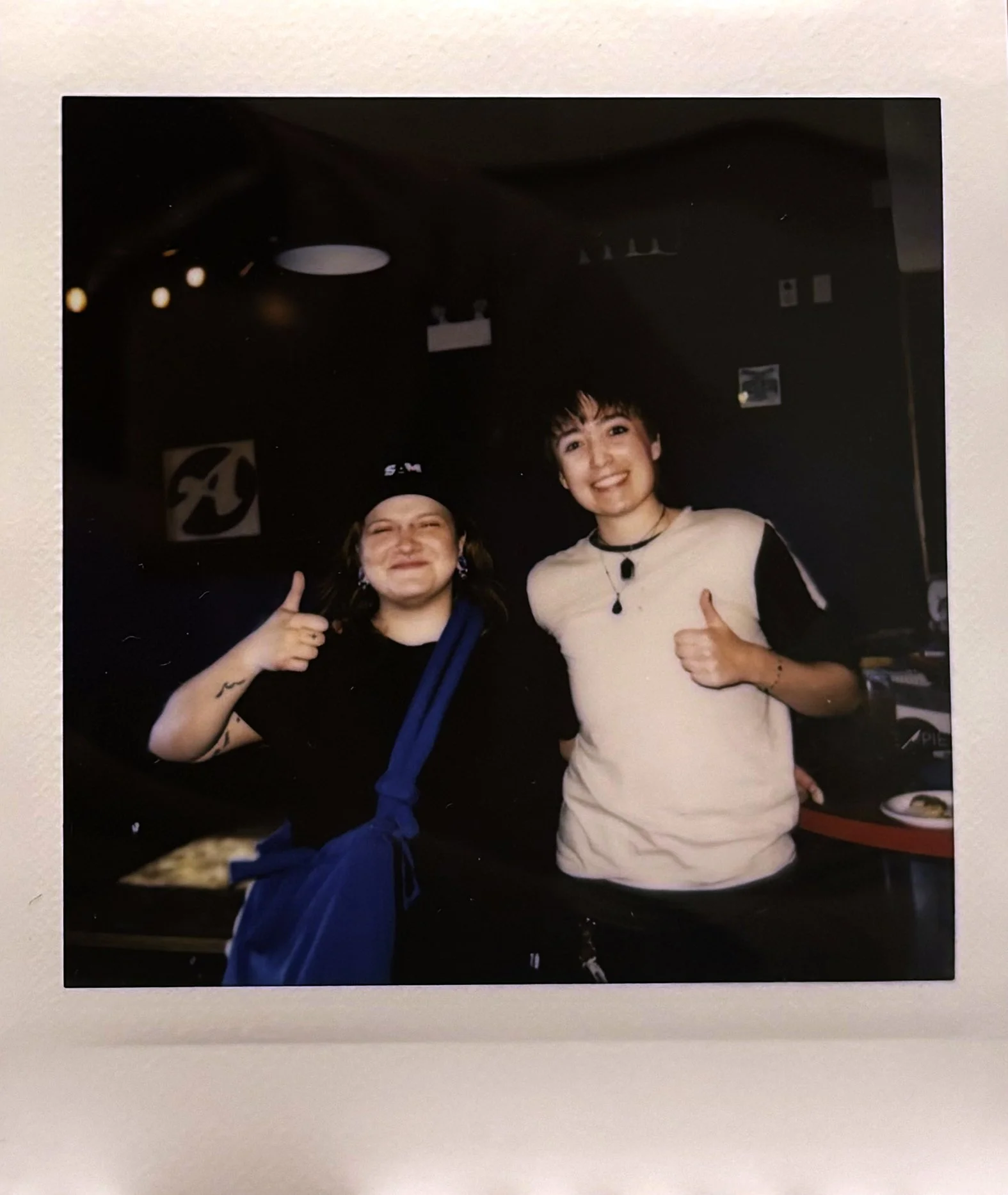 Two smiling women smiling with thumbs up, one with short dark hair and the other with dark hair covered with a black cap, in an indoor setting with dark walls and some artwork or posters in the background.