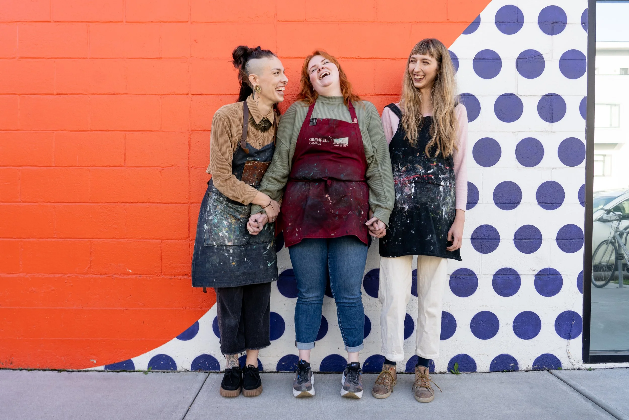 Three women laughing and holding hands in front of a colorful mural with orange and white sections and blue polka dots.