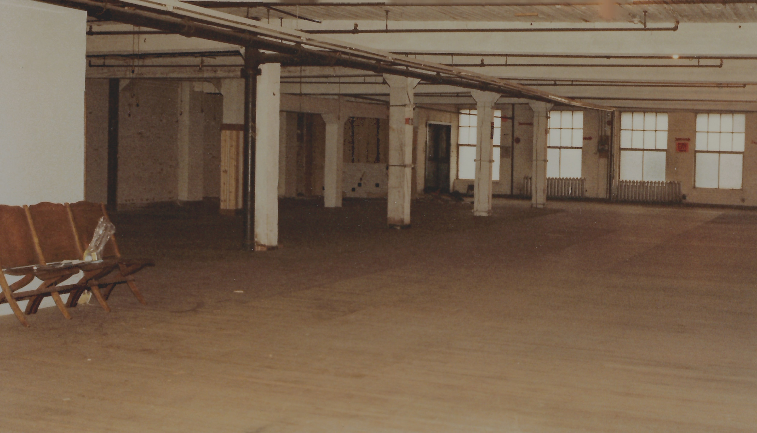 Empty large industrial space with high ceilings, exposed pipes, white support columns, and large windows along the back wall. Brown wooden chairs are lined up along the left side, with some objects on them. The floor appears to be unfinished or dusty.